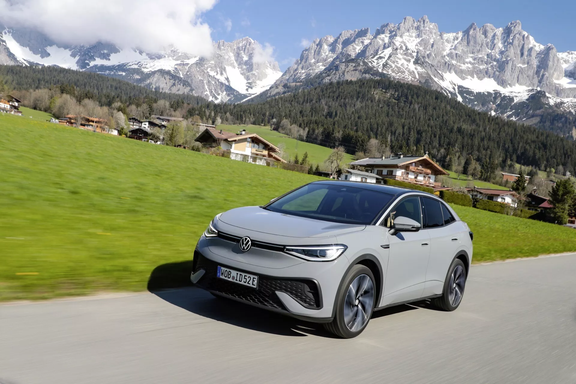 Obraz elegantného strieborného crossoveru SUV Volkswagen zachyteného v jasný slnečný deň s horskou krajinou a jasnou oblohou v pozadí. Auto je vidieť z nízkeho uhla, ktorý ukazuje prednú ľavú stranu, s ostrými svetlometmi, úzkou mriežkou, viackôlovými zliatinovými kolesami a elegantnými líniami naznačujúcimi moderný, možno elektrický alebo hybridný dizajn. Auto je na vydláždenej ceste a zdá sa, že sa pohybuje proti pokojnému vidieckemu pozadiu.