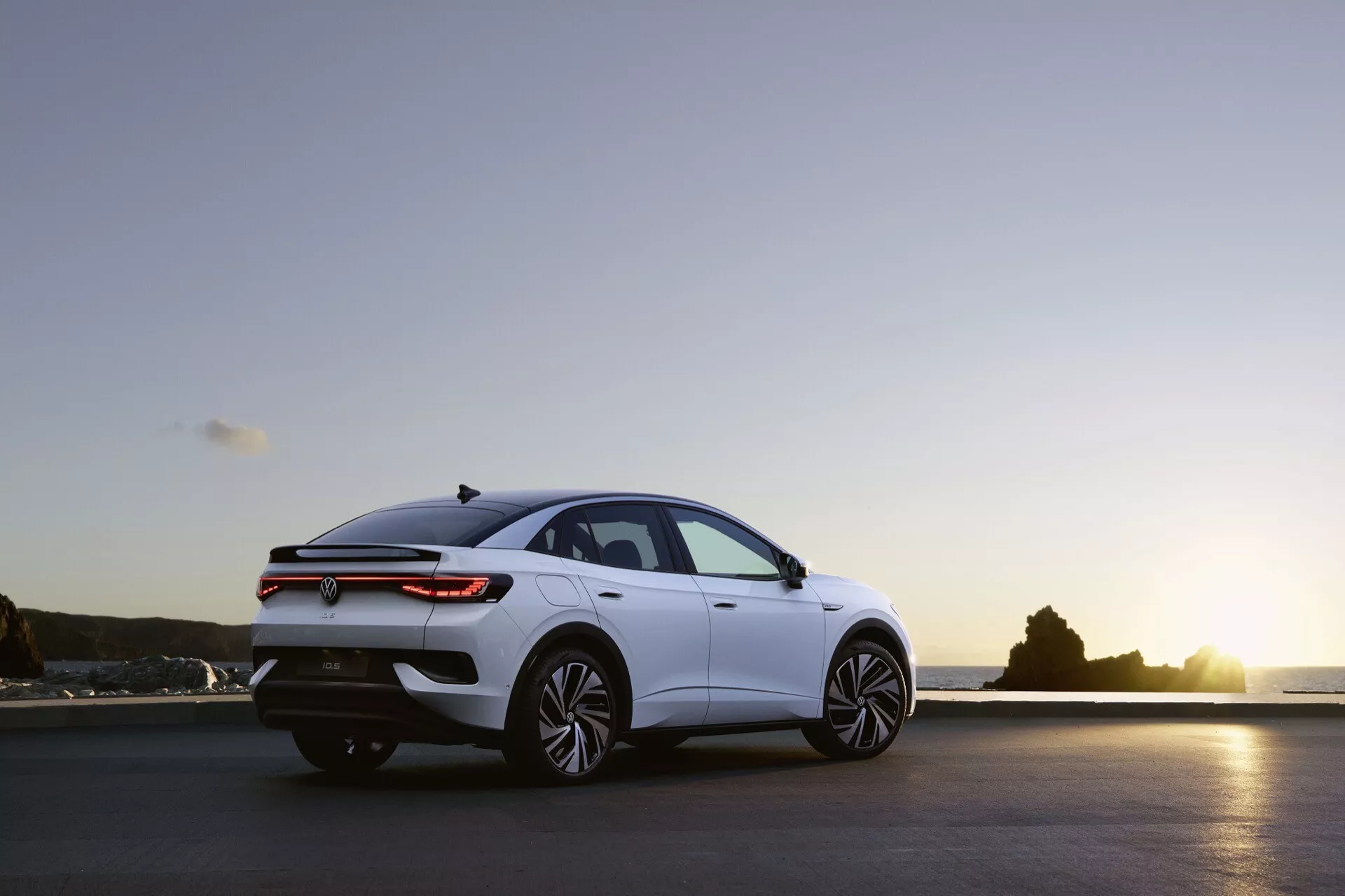 Bočný profil sedanu VW ID.5 prvej generácie z roku 2022 v exteriéri počas skorého večera so jemným osvetlením. Nízky uhol snímky zvýrazňuje aerodynamické línie, elegantný dizajn a štýlové viac-lúčové hliníkové disky. Moderné prvky zahŕňajú ostré svetlomety, štíhle zadné svetlá a subtilný spojler na veku batožinového priestoru. Rozsiahle nebo s farbami západu slnka a prírodné pozadie zdôrazňuje modernú, luxusnú estetiku vozidla.