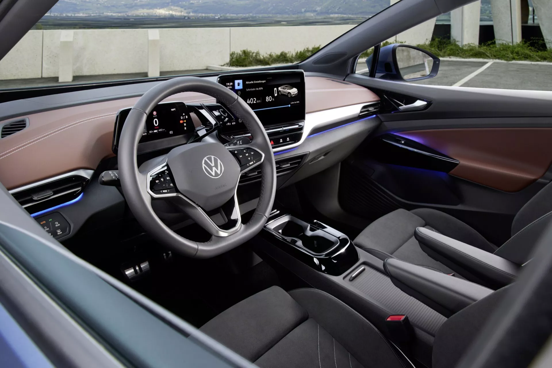Pohľad do interiéru Volkswagen ID.5 2022 (1. generácia) z pohľadu vodiča, ktorý zobrazuje multifunkčný volant s logom Volkswagen, digitálny prístrojový panel a centrálnu infotainment dotykovú obrazovku. Moderná stredová konzola obsahuje elektronickú radiacu páku a pravdepodobne aj volič jazdných režimov, všetko zvýraznené ambientným osvetlením. Dvojfarebné sedadlá z kože a semišového materiálu a vysoko kvalitné materiály dverových panelov zdôrazňujú luxusný a sofistikovaný dizajn.