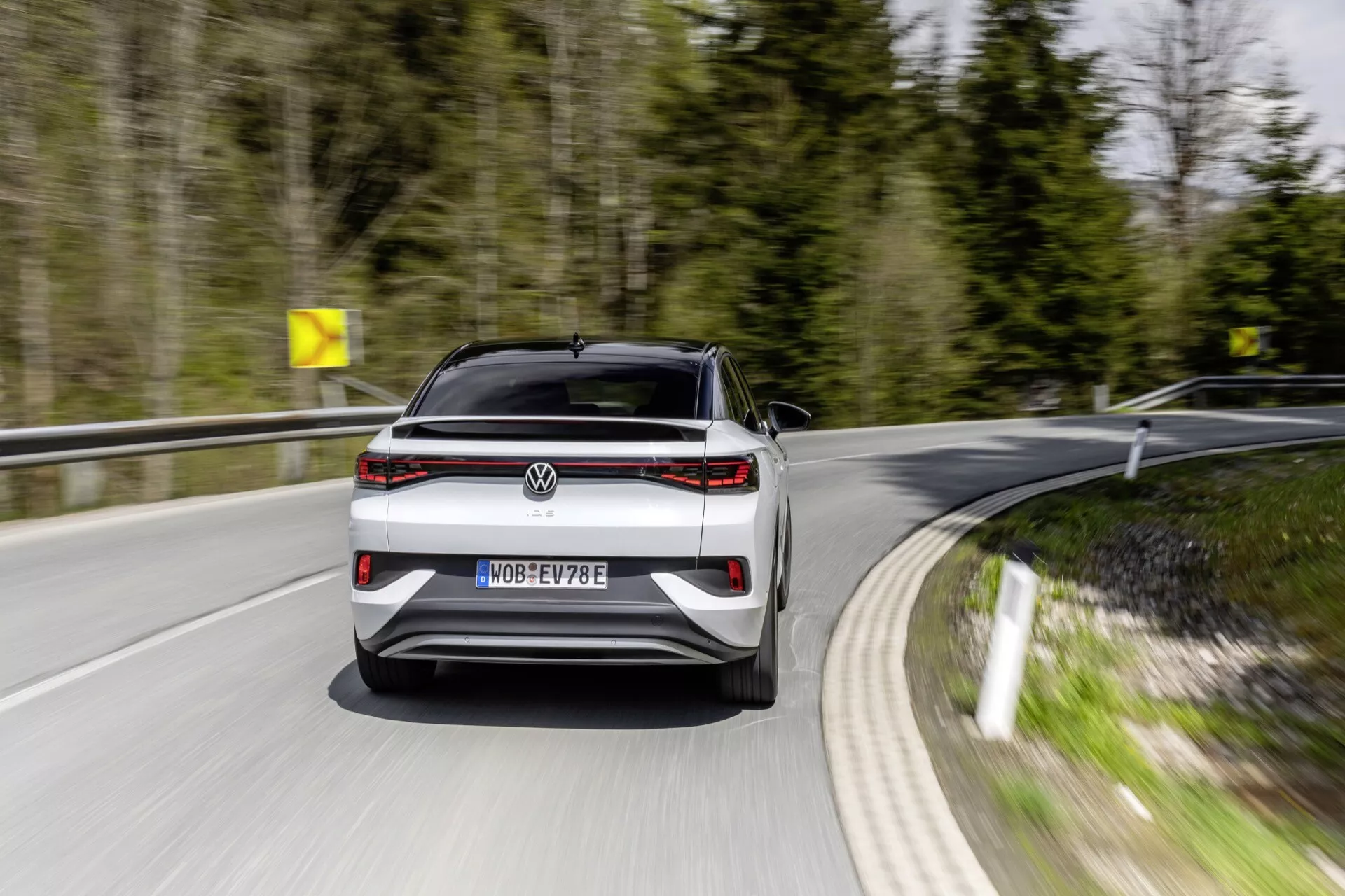Dynamický pohľad na zadnú časť pohyblivého VW ID5 2022 (1. generácia) na zakrivenej ceste, zachytený z nízko-strednej výšky, so zameraním na moderné dizajnové prvky ako výrazný horizontálny svetelný pás, rozoznateľný emblém a označenie modelu. Evidenčné číslo naznačuje, že ide o elektrické vozidlo s označením 'E'. Viditeľné parkovacie senzory a absencia výfukových potrubí naznačujú pokročilé funkcie, ako je technológia parkovacej asistencie a elektrický pohon.