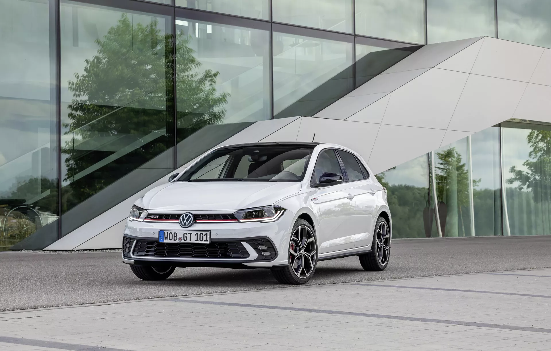 Moderný hatchback v lesklom bielom odtieni umiestnený na ľavej strane rámu, fotografovaný z nízkeho, trojštvrťového predného uhla. Elegantný a športový exteriér zahŕňa naklonenú kapotu, dynamické línie, ostré LED svetlomety, výraznú prednú mriežku, integrované hmlové svetlá, výkonné nasávacie otvory a viacpaprskové zliatinové kolesá s dvojfarebným povrchom. Auto sa nachádza v mestskom prostredí s modernou budovou so šikmými dizajnmi a reflexným sklom.
