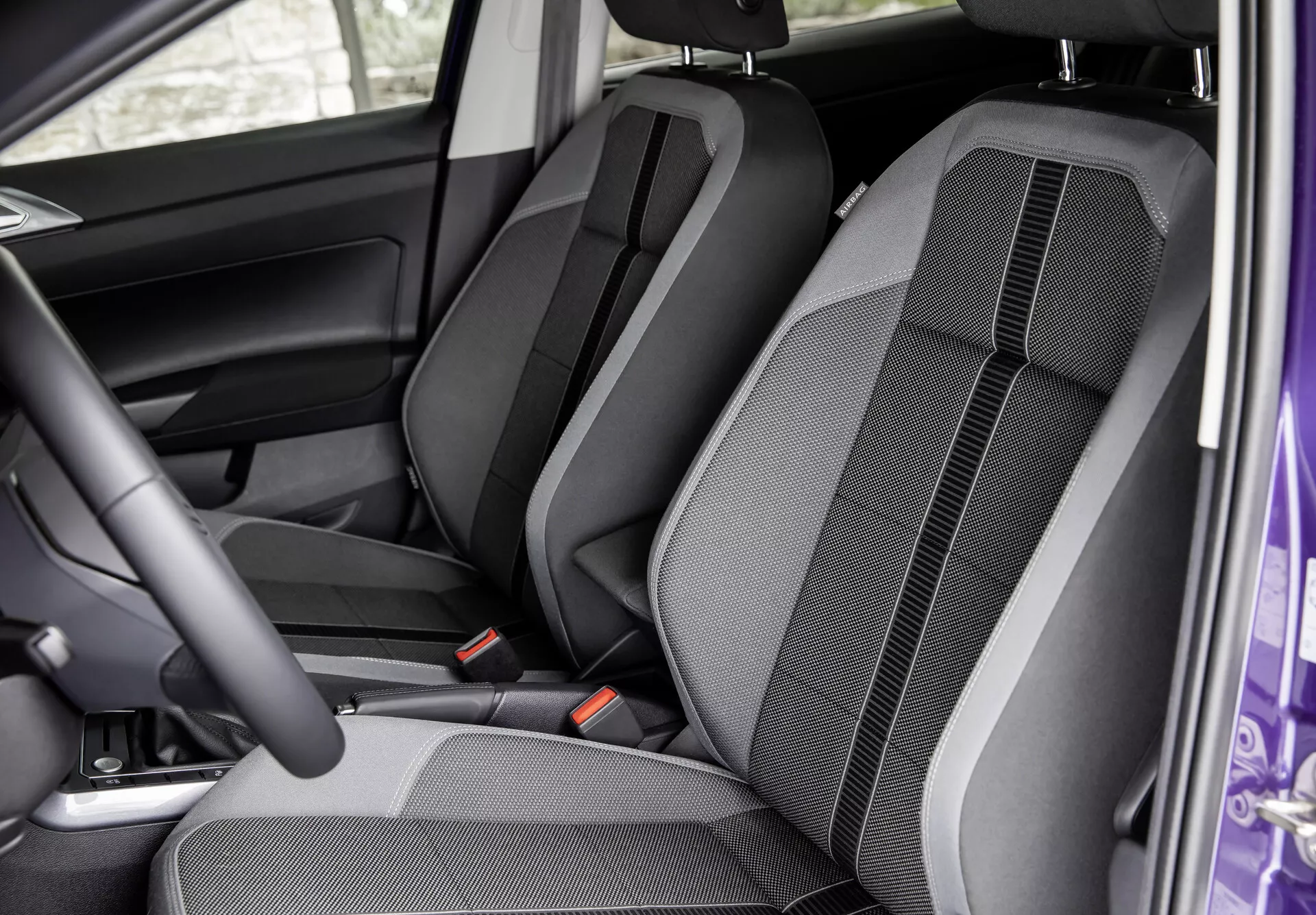 Interiér VW Polo 2021 (VI-facelift) z pohľadu otvorených predných dverí na sedadlá predných a zadných sedadiel. Zobrazená je konfigurácia sedadiel, dizajn látky, prešívanie a detaily tvarovania. Viditeľné sú výškovo nastaviteľné opierky hlavy, bezpečnostné pásy a čiastočný pohľad na dverný panel pri vodičovi, zdôrazňujúci komfort a interiérový dizajn.