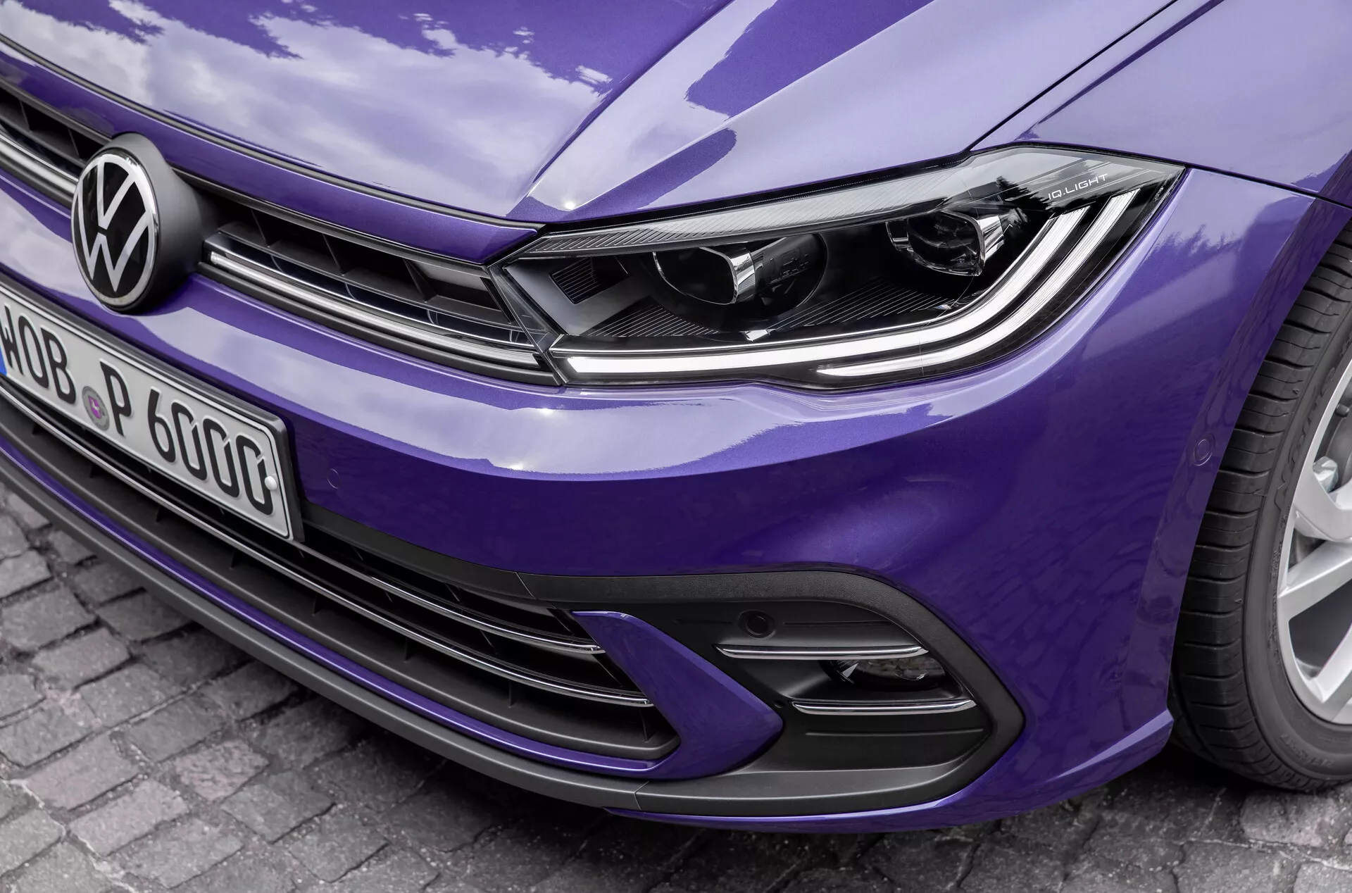 Detailný, uhlový predný pohľad na modrý Volkswagen Polo 2021 VI-facelift, ktorý zobrazuje moderný dizajn svetlometov s integrovanými LED dennými svetlami, výrazné logo Volkswagen na horizontálnych líniách mriežky a športový nárazník s prívodmi vzduchu a dizajnom včelieho plástu. Detaily zvýrazňujú elegantné línie kapoty a lesklý povrch laku, čo naznačuje pokročilú osvetľovaciu technológiu a potenciálne systémy asistencie vodiča.