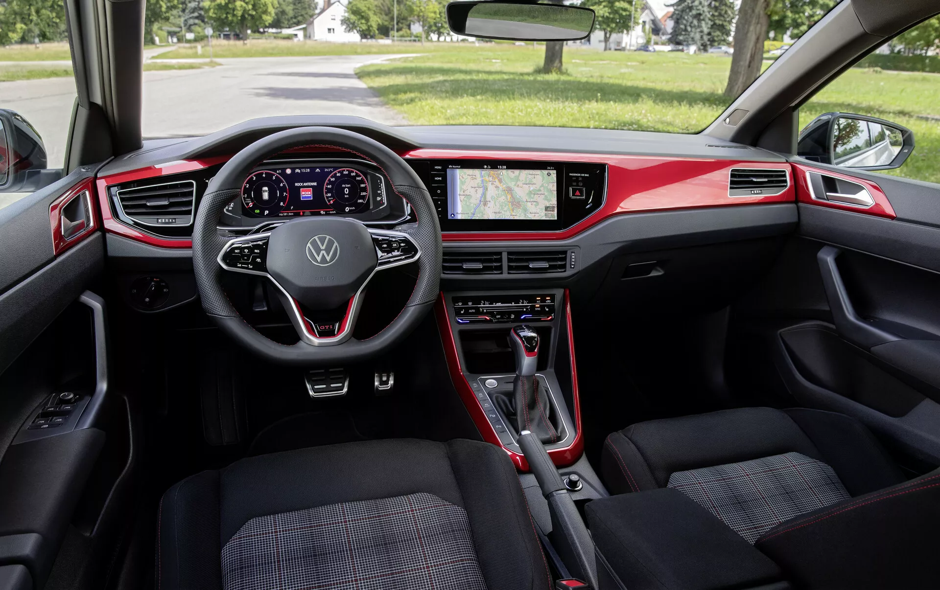 Pohľad do interiéru VW Polo 2021 (VI-facelift) z perspektívy zadného sedadla. Širokouhlý záber zachytáva palubnú dosku, predné sedadlá a spätné zrkadlo. Medzi výrazné prvky patrí moderný multifunkčný volant s logom Volkswagen, plne digitálny prístrojový štít, veľký dotykový infotainment displej zobrazujúci GPS navigáciu a fyzické ovládanie klimatizácie. Prevažne čierny interiér s červenými akcentmi zdôrazňuje športový dizajn. Predné sedadlá majú vzorované čalúnenie a radiaca páka naznačuje automatickú prevodovku. Celkovo interiér spája funkčnosť s moderným dizajnom a pokročilou technológiou.