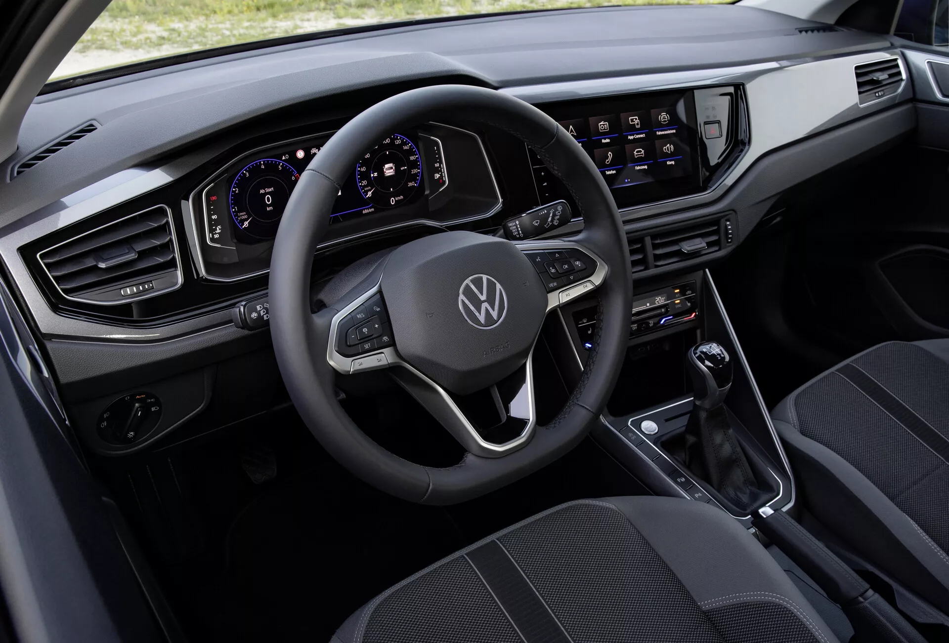 Pohľad z výšky na interiér VW Polo 2021 (VI-facelift), ktorý ukazuje multifunkčný kožený volant s plochým spodkom zdobený logom Volkswagen, plne digitálny a prispôsobiteľný prístrojový štít, veľký dotykový infotainment systém, modernú vodorovnú palubnú dosku s elegantnými, hranatými vetracími otvormi, minimalistickú stredovú konzolu s radiacou pákou pre automatickú prevodovku a kvalitné látkové sedadlá so vzorovaným dizajnom.