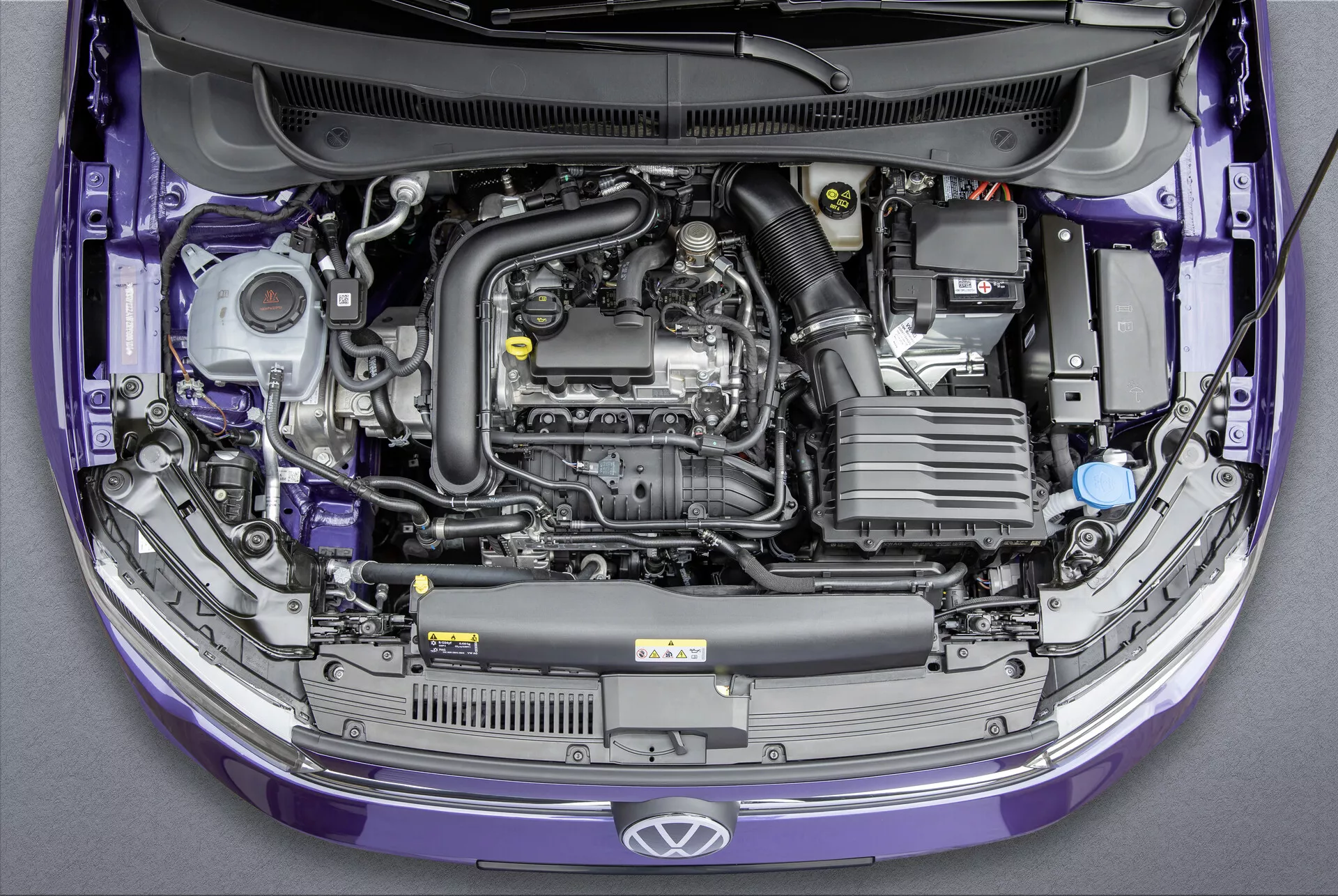 Pohľad zhora na Volkswagen Polo 2021 (VI-Facelift) so zdvihnutou kapotou, ktorá odhaľuje priečne umiestnený motor a rôzne komponenty v motorovom priestore. Obrázok zvýrazňuje plastový kryt motora, batériu, systém nasávania vzduchu, nádržku chladiacej kvapaliny s modrým viečkom, rôzne nádoby na kvapaliny, súčasti motora s kovovým a gumovým potrubím a skrinku s poistkami. Karoséria auta je fialová a v štandardnom továrenskom stave. Ideálne pre kategórie týkajúce sa automobilových motorov, údržby vozidiel, automobilového inžinierstva a výroby automobilov.