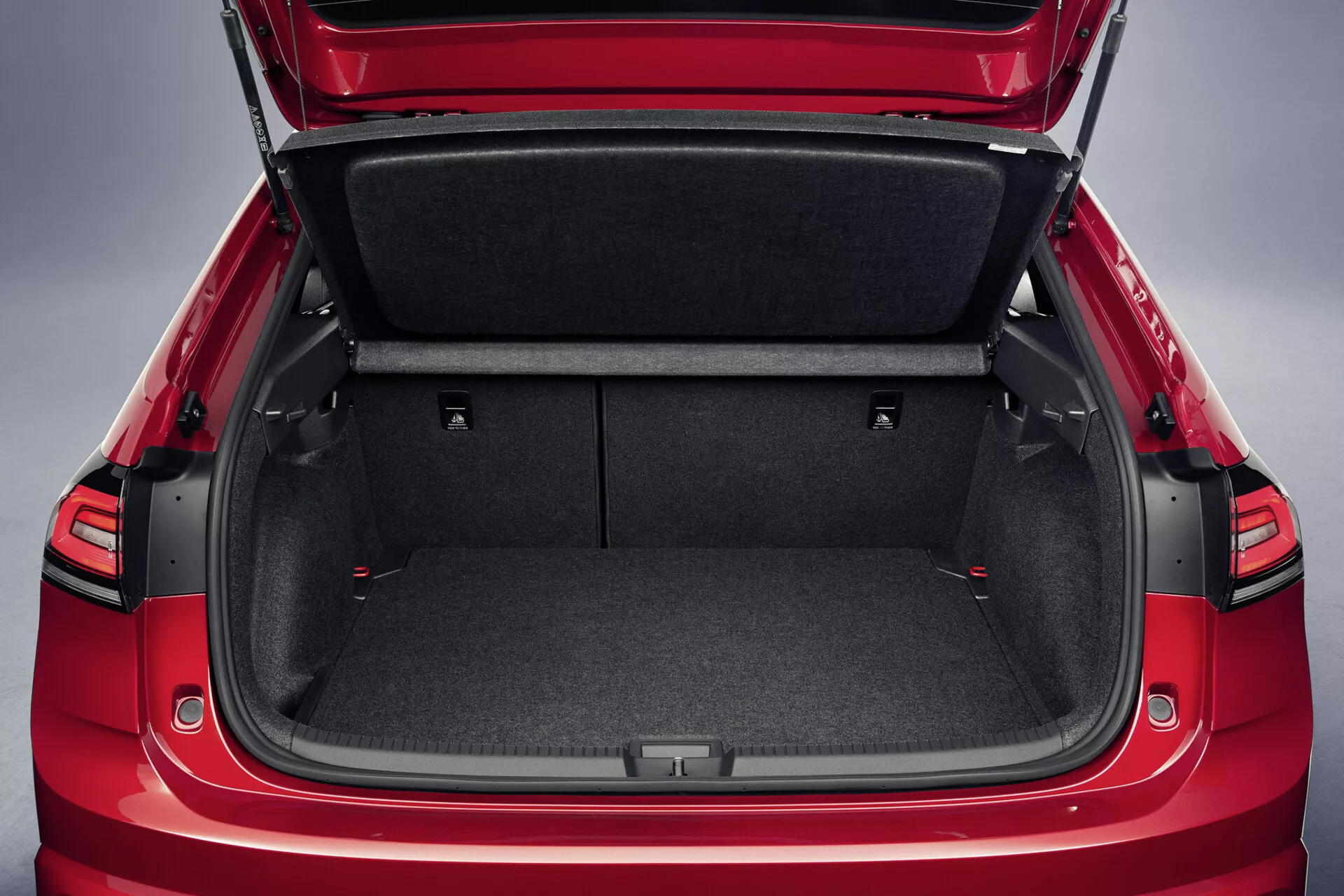 Detailný pohľad na červený exteriér a priestranný interiér batožinového priestoru VW Taigo 2021 s odolnou kobercovou výstelkou, sklopnými zadnými sedadlami s červenými ťahacími popruhmi, zasúvateľnou krytkou nákladu a úložnými priestormi