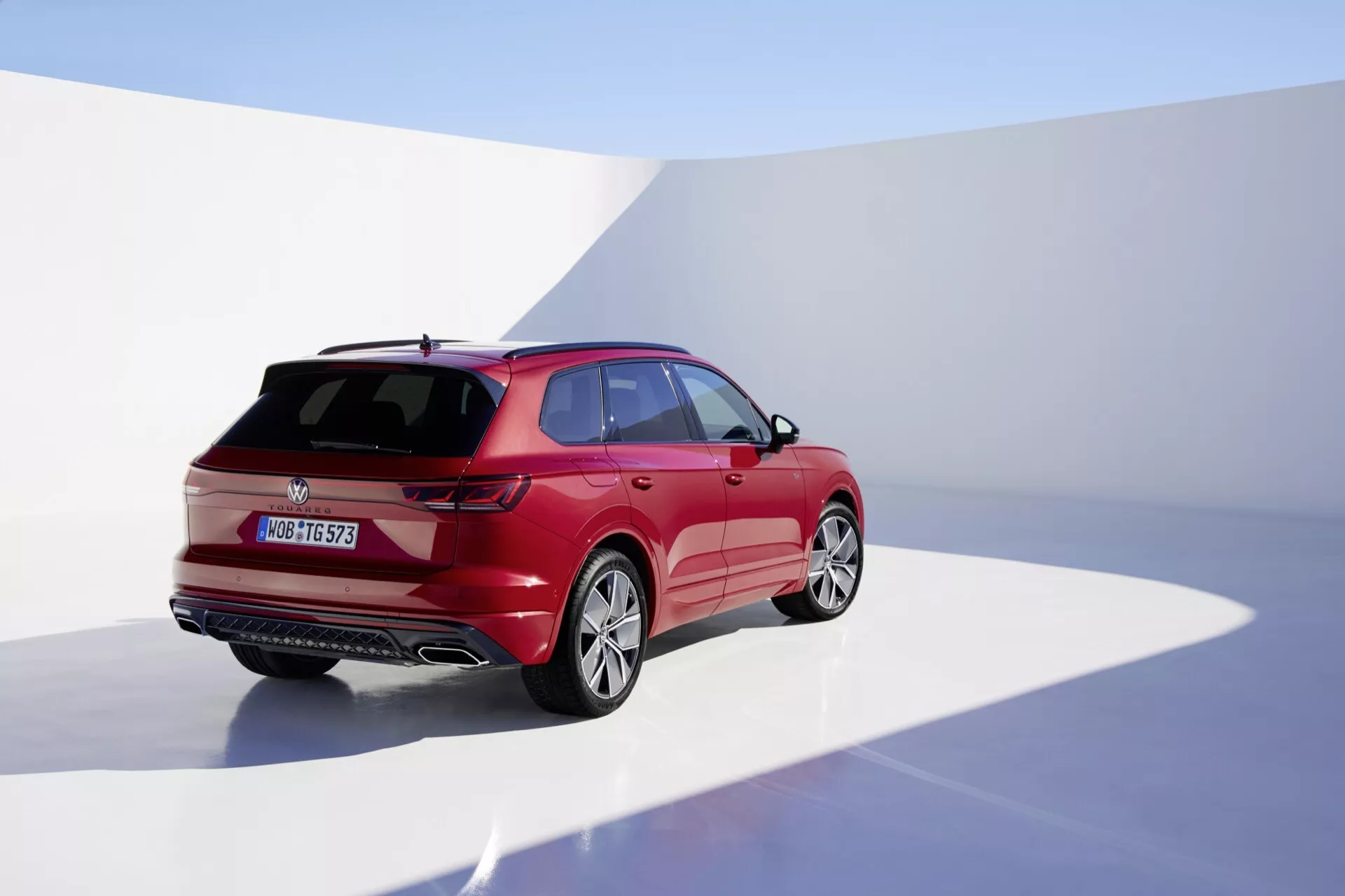 Model VW Touareg III facelift 2023 sfotografovaný z pohľadu zo zadného trojštvrtinového uhla zdôrazňujúci pravú stranu, v štúdiu s plynulým pozadím, predvádzajúci jeho lesklý červený povrch, aerodynamický dizajn s ostrými karosárskymi líniemi, LED zadné svetlá, zadný stierač, strešný spoiler a zliatinové kolesá.