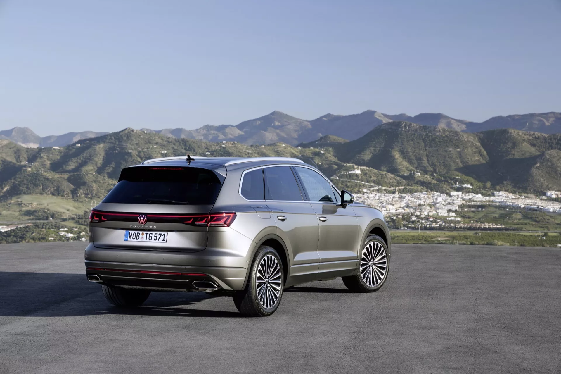 SUV VW Touareg 2023 (III Facelift) zaparkované vonku s malebným pozadím kopcov a malého mesta alebo predmestia, ktoré ukazuje pravý zadný štvrťrok a bočný profil, s dôrazom na zadné svetlá, líniu strechy a línie obrysu karosérie. Funkcie zahŕňajú veľké zliatinové kolesá, panoramatické strešné okno, LED zadné svetlá a prémiové prvky off-road štýlingu.