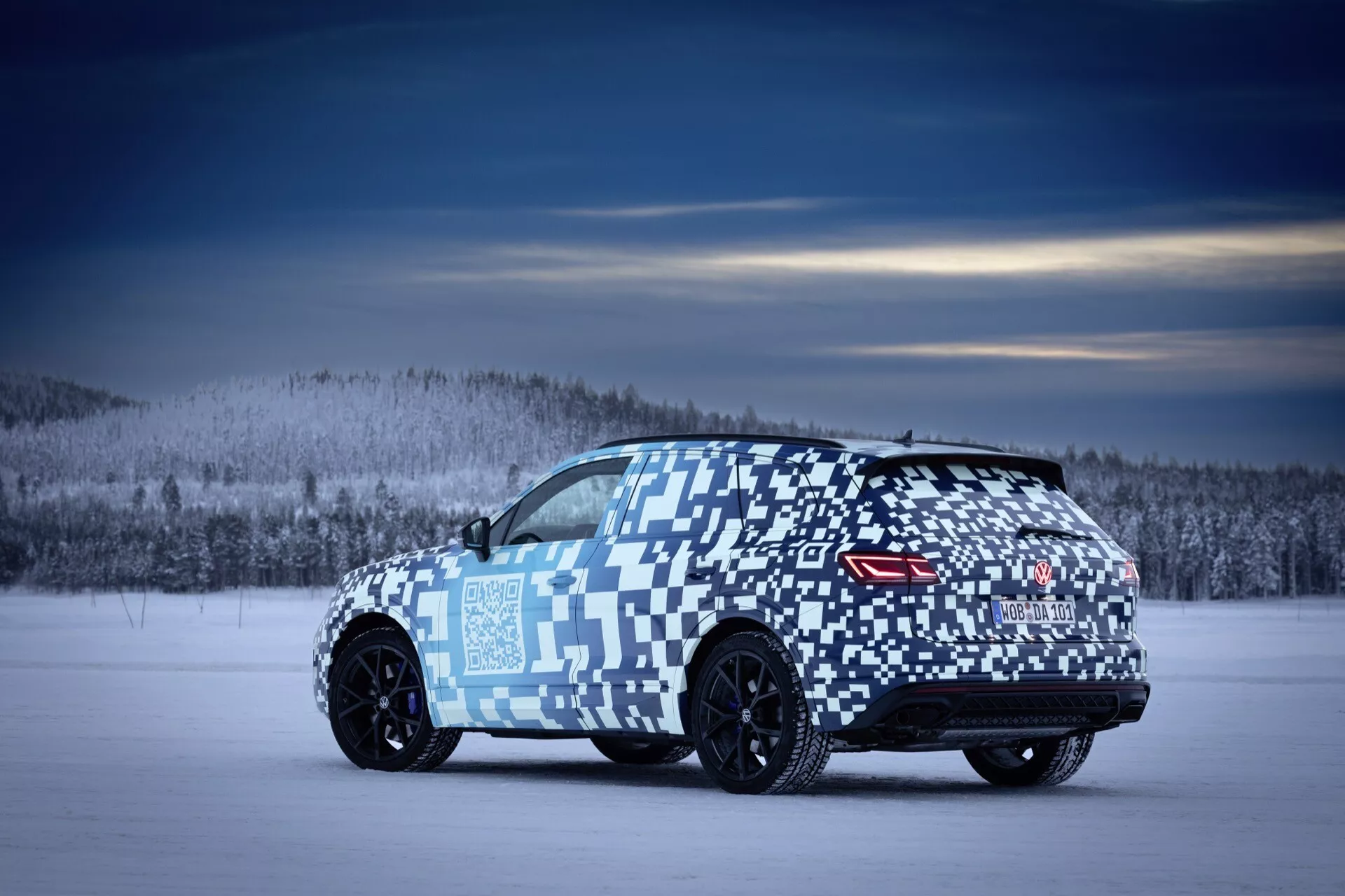 VW Touareg (III-facelift) 2023 zozadu z uhla troch štvrtín na zasneženom teréne s maskovacím obalom, čiernymi ľahkými kolesami, zadnými LED svetlami a spojlerom naznačujúcim orientáciu na výkon