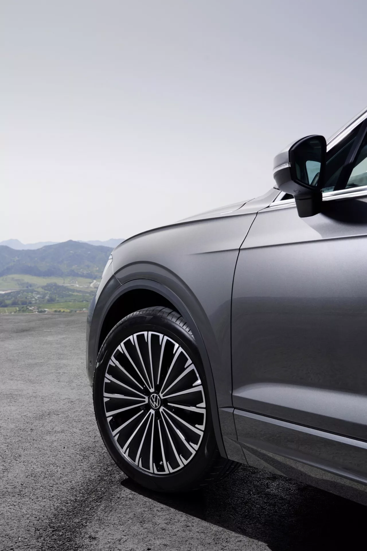 Detailný pohľad na prednú časť vozidla na strane vodiča Volkswagen Touareg 2023 (III Facelift) s veľkým, štýlovým zliatinovým kolesom s komplexným dizajnom viacerých špiciek, odrážajúcim kvalitu vozidla prémiovej triedy na pozadí horskej krajiny.