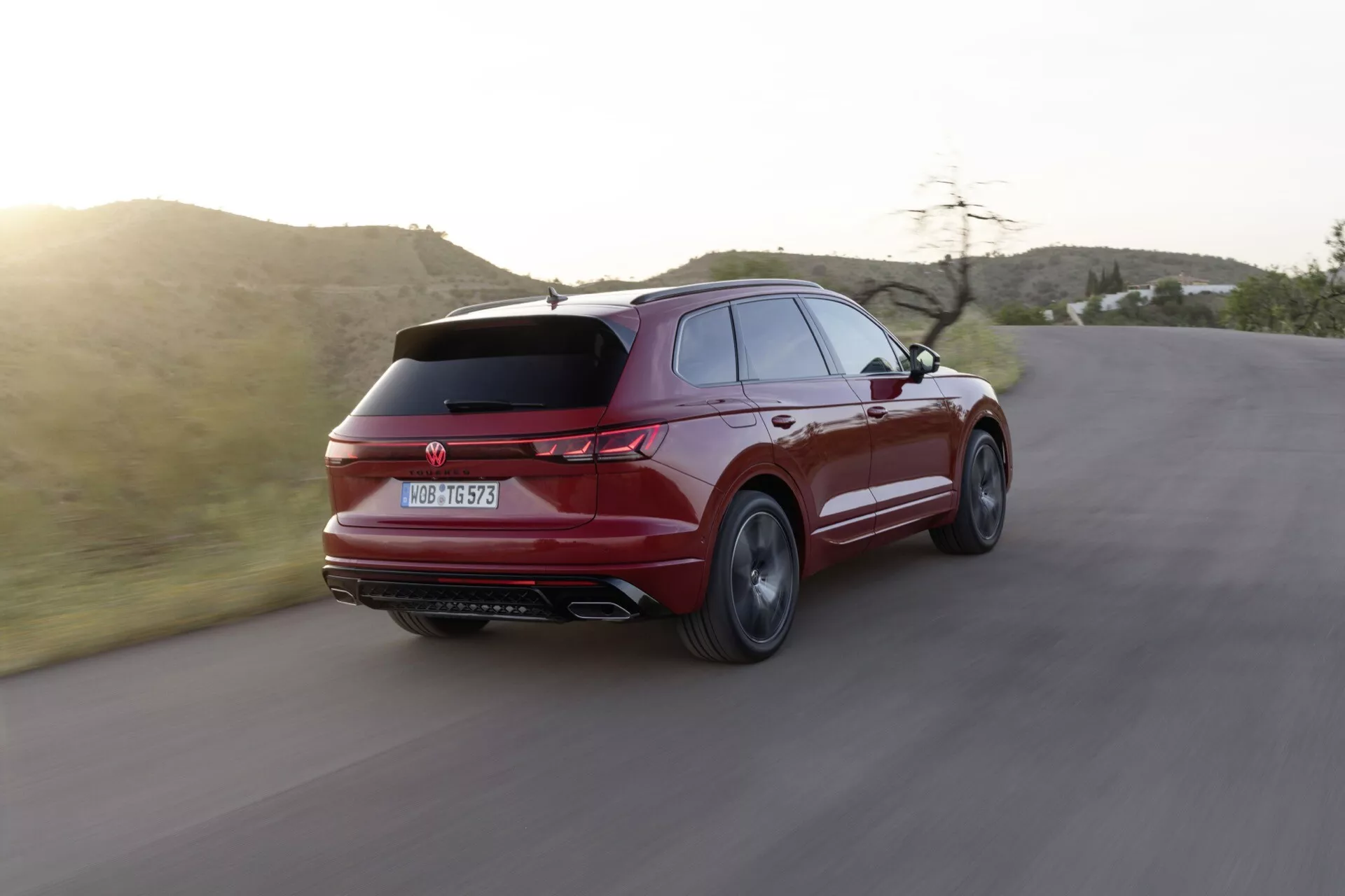 Facelift Volkswagen Touareg III 2023 zachytený v dynamickom trojštvrťovom zadnom pohľade na zákrute, zdôrazňujúci jeho športovo elegantný dizajn, štíhly zadný spojler, charakteristické zadné svetlá, dvojité výfukové trubky a viacramenné zliatinové kolesá na pozadí polosuché krajiny počas zlatej hodiny