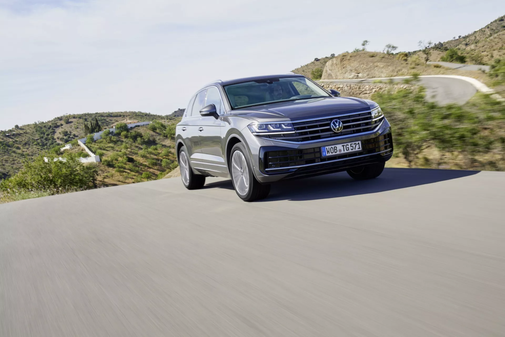 SUV Volkswagen Touareg III Facelift z roku 2023 v pohybe na asfaltovej ceste, s výraznou prednou mriežkou, LED svetlometmi a elegantným obrysom kapoty na pozadí prírodnej krajiny, zdôrazňujúcim moderný dizajn a automobilovú technológiu.