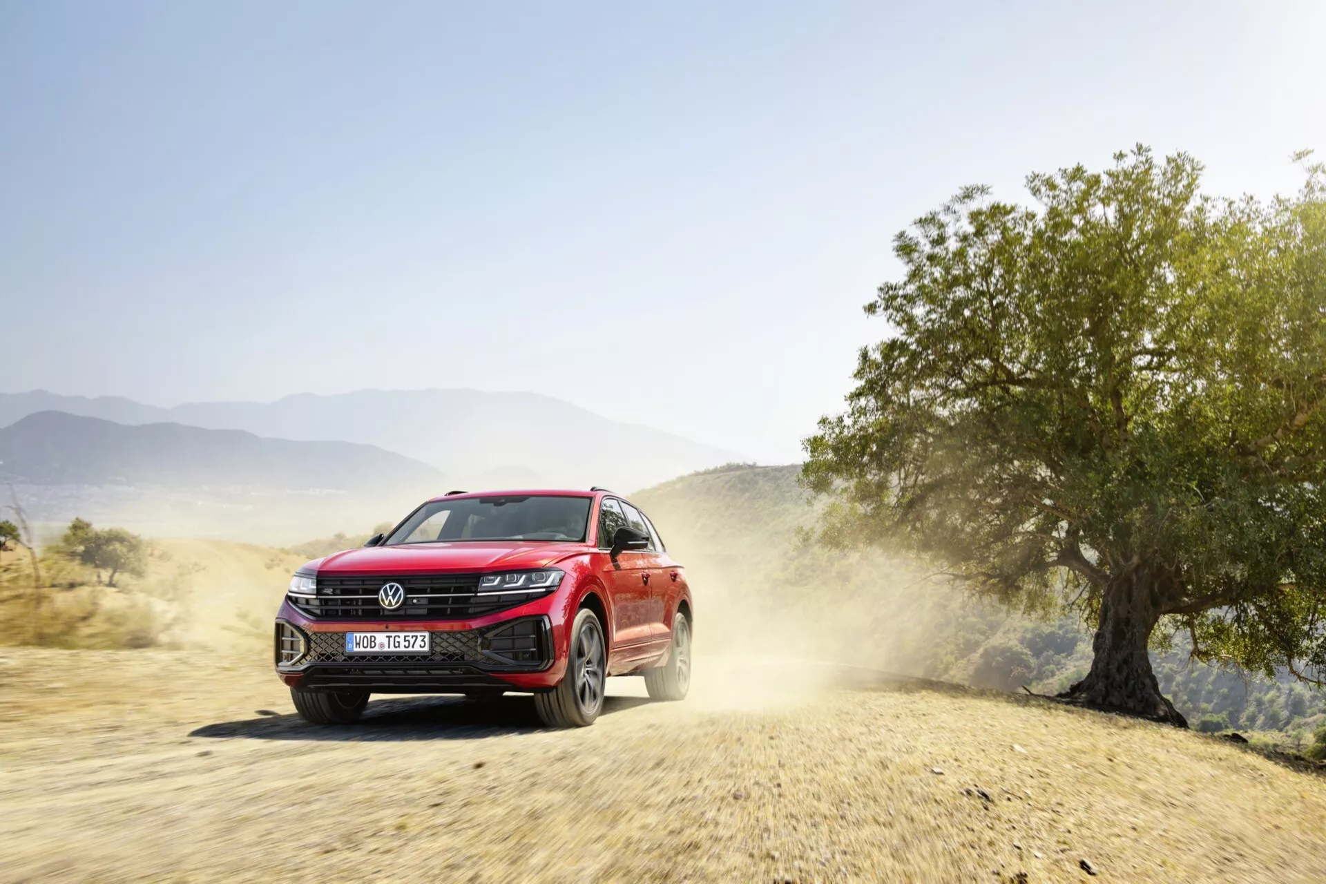 Červené SUV VW Touareg 2023 v dynamickom pohybe na neupravenom ceste so stopou prachu, ukazujúce prednú fasádu, LED svetlomety, mriežku s potenciálnymi senzormi asistenčného systému vodiča a zliatinové kolesá, naznačujúce schopnosti v teréne a moderné technologické funkcie