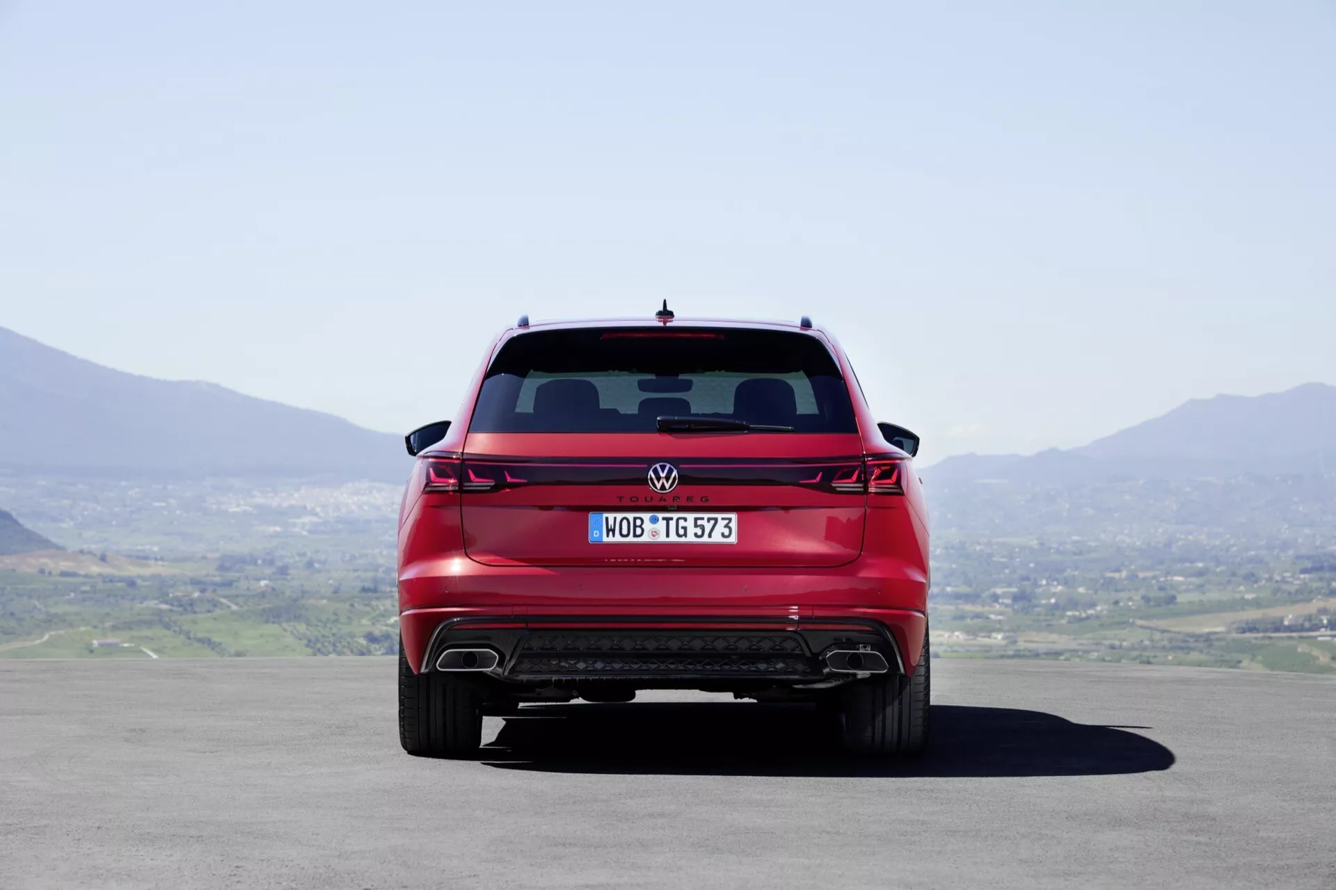 Zadný pohľad na Volkswagen Touareg 2023 ukazuje celú šírku, moderné zadné svetlá, zadný difúzor a dvojité výfukové koncovky, pod prírodným denným svetlom s čistou oblohou a kopcovitým pozadím krajiny