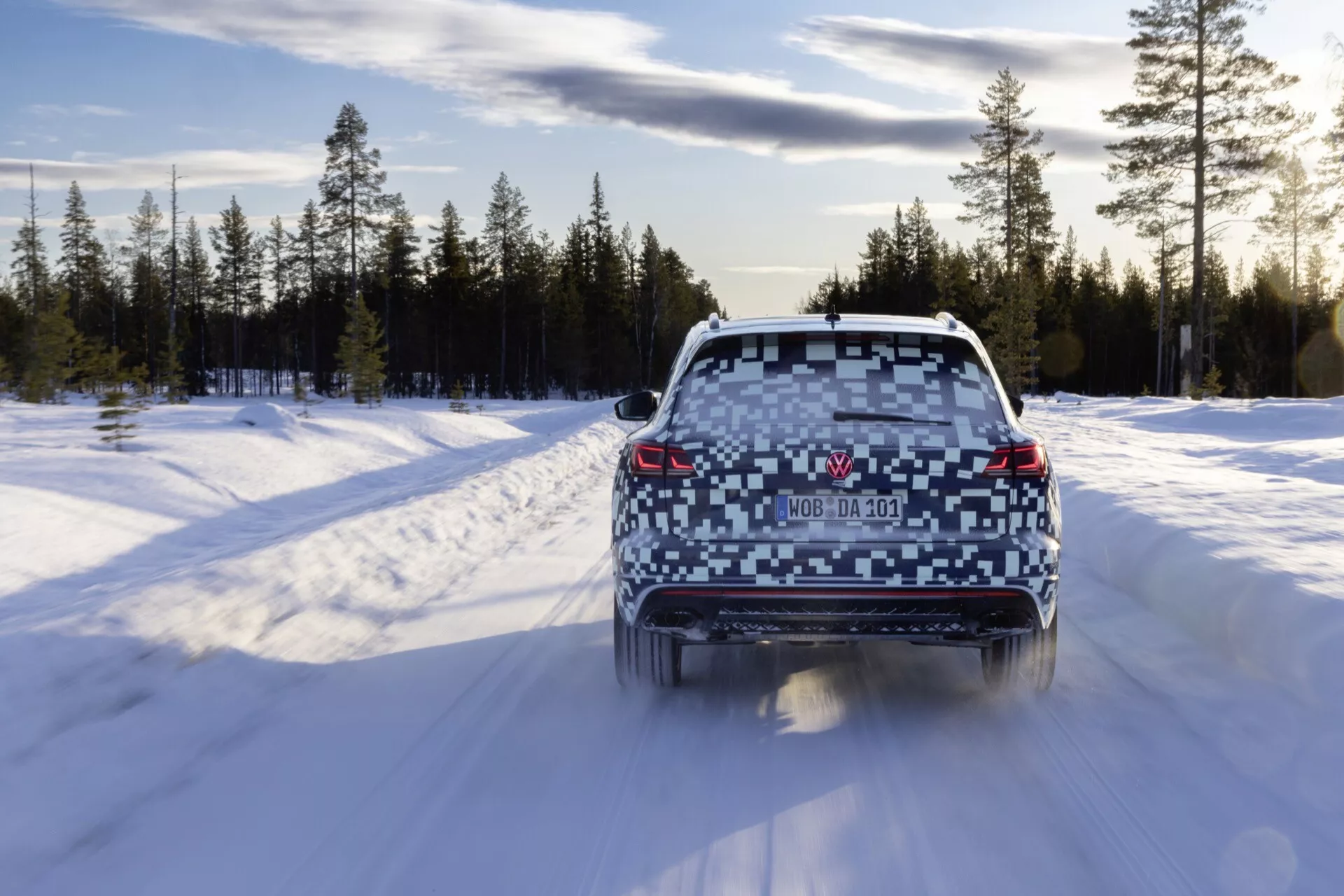 Zadný pohľad na Volkswagen Touareg 2023 (III-Facelift) idúc na zasneženej ceste v zimnom lese, ukazujúci svietiace zadné svetlá, čisté zadné okno, dvojité výfuky a strešný spoiler.