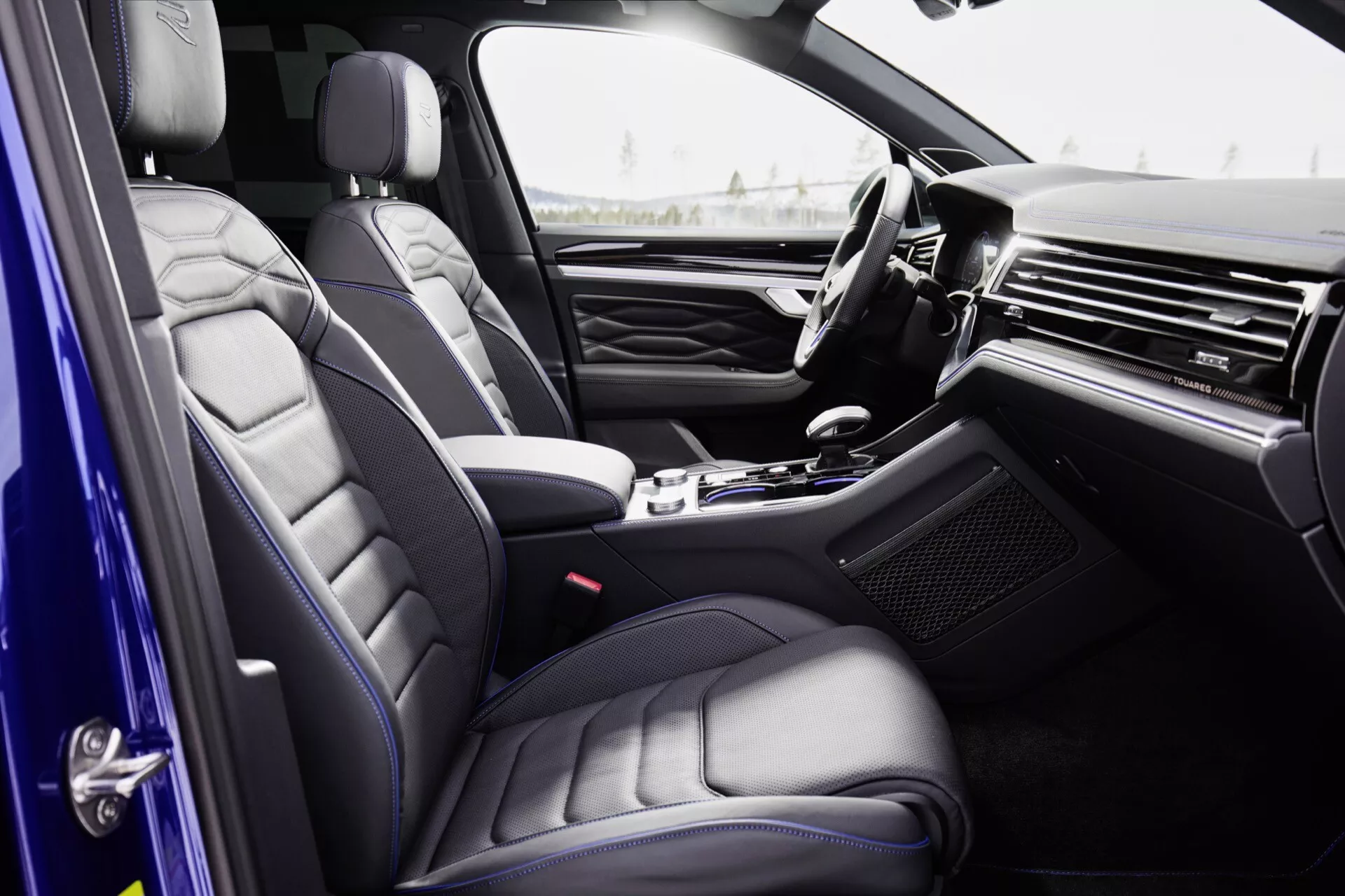 Interiér Volkswagen Touareg 2023 (III facelift) z pohľadu cestujúceho, s dôrazom na sedadlá z prémiovej kože s prešívaným dizajnom, elegantnú palubnú dosku s kovovým alebo drevným obložením, luxusnú stredovú konzolu s voličom automatickej prevodovky, dverový panel s vysokou úrovňou výbavy a čiastočne viditeľný volant s integrovanými ovládačmi.