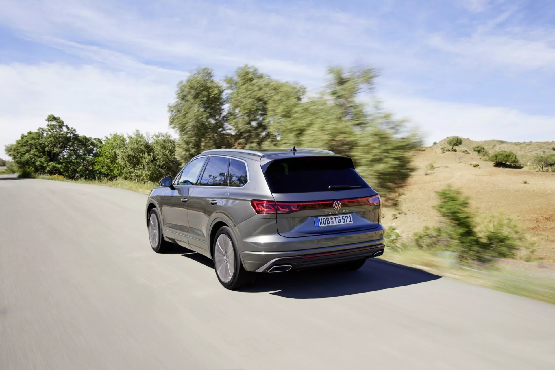 Facelift VW Touareg III z roku 2023 zobrazený z pohľadu zo zadného trojštvrte z uhla pri pohybe počas dňa za jasného počasia, predvádzajúci LED zadné svetlá, štýlový design kombi s mierne sa zvažujúcou strechou a strešným spoilerom, čiastočný pohľad na dizajn zliatinového kolesa a metalický lak.