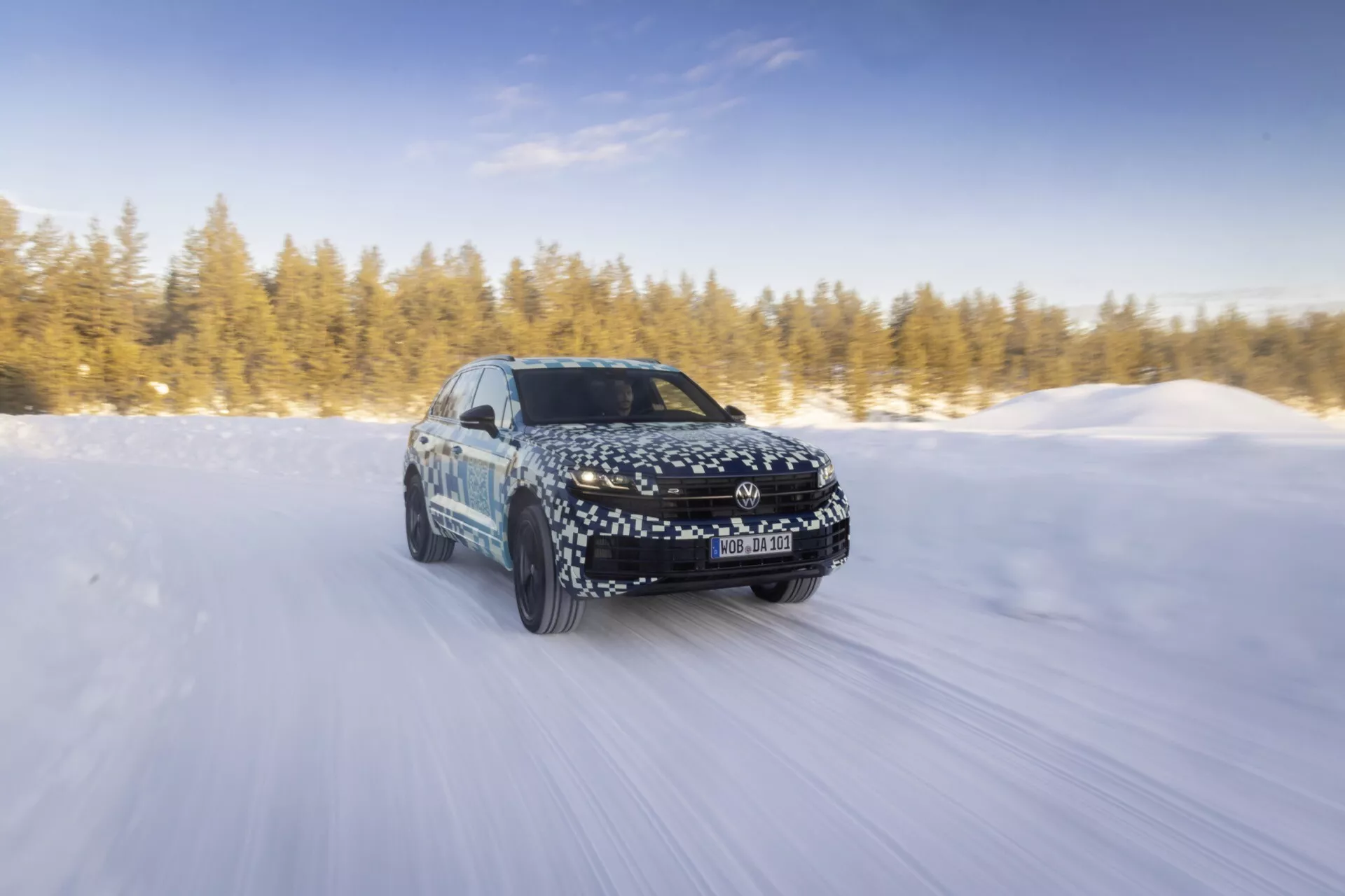 Model Volkswagen Touareg III facelift z roku 2023 dynamicky jazdiaci po zasneženej ceste na slnku, ukazujúci pohyb vpred, LED svetlomety, zliatinové kolesá a jeho aerodynamický dizajn s maskovacím obalom.