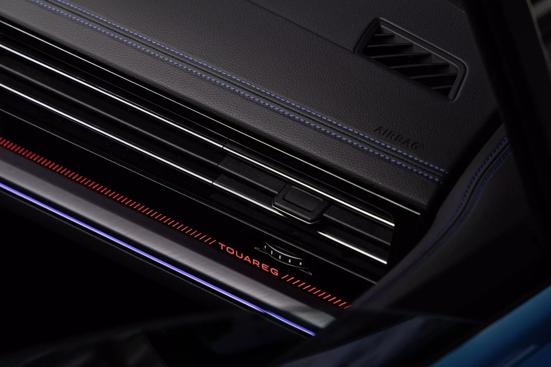Detailný pohľad na interiér Volkswagen Touareg facelift 2023 so zameraním na dizajn prístrojovej dosky, výrazný nápis 'Touareg' s ambientným osvetlením, ventilačné mriežky so striebornými akcentmi a atmosférický červený svetelný pruh pre pocit luxusu