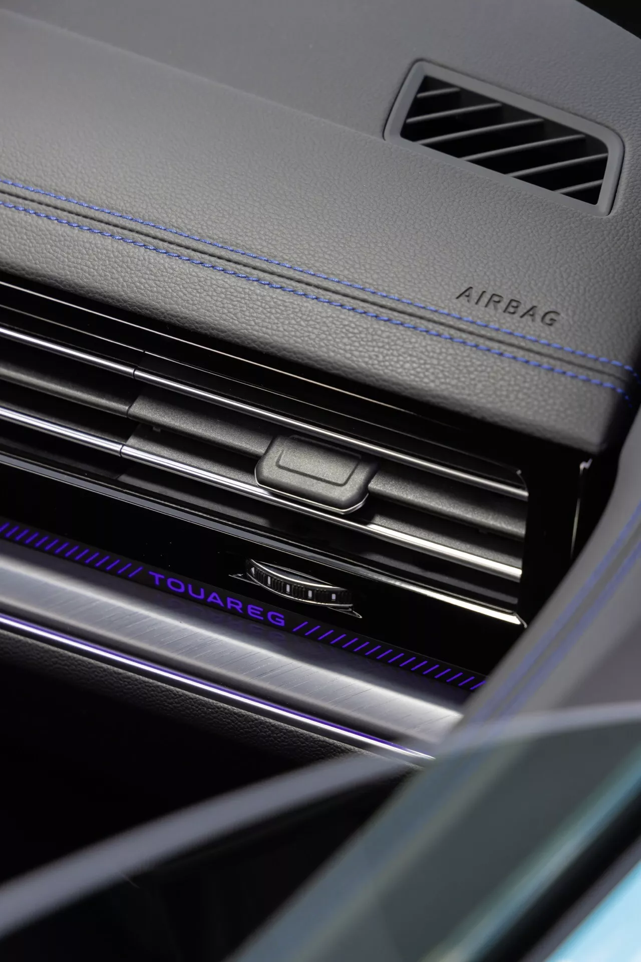 Zblízka uhlový pohľad na palubnú dosku na strane spolujazdca Volkswagen Touareg 2023 (III-Facelift) s zdôraznením indikácie AIRBAG, chrómovaných výustiek klimatizácie so stredovým ovládacím gombíkom, osvetleným názvom 'TOUAREG', luxusnými materiálmi s kontrastným šitím a premyslenými prvkami interiérového dizajnu v prostredí s nízkym osvetlením zdôrazňujúcimi pokročilú technológiu vnútorného osvetlenia vozidla a bezpečnostné funkcie.