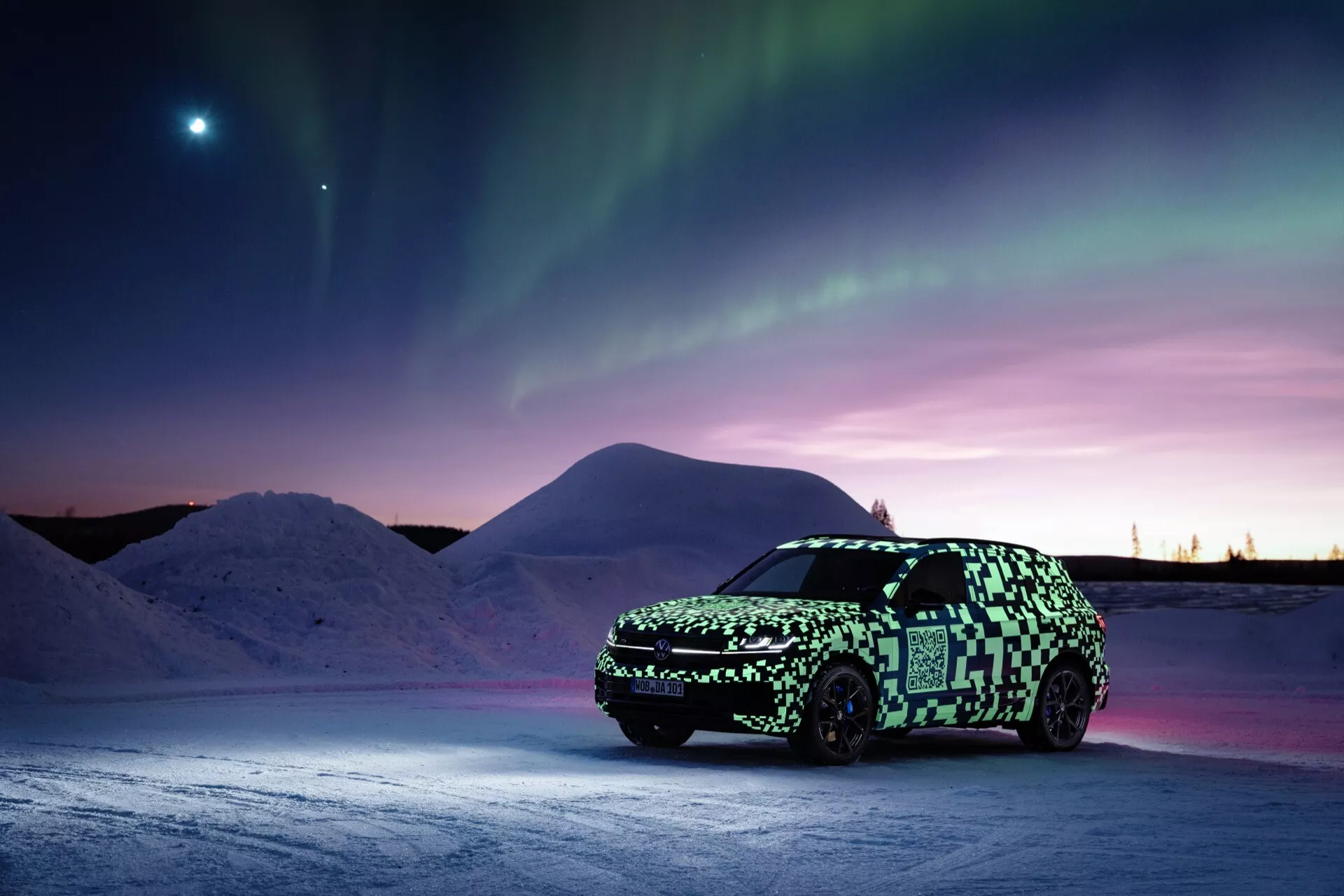 Prototyp VW Touareg (III-facelift) v maskovacej fólie na zasneženom povrchu pod polárnou žiarou s jasným nebeským telesom v súmrakovom nebi, zdôrazňujúci pokročilé osvetlenie a potenciálnu elektrickú alebo hybridnú technológiu.