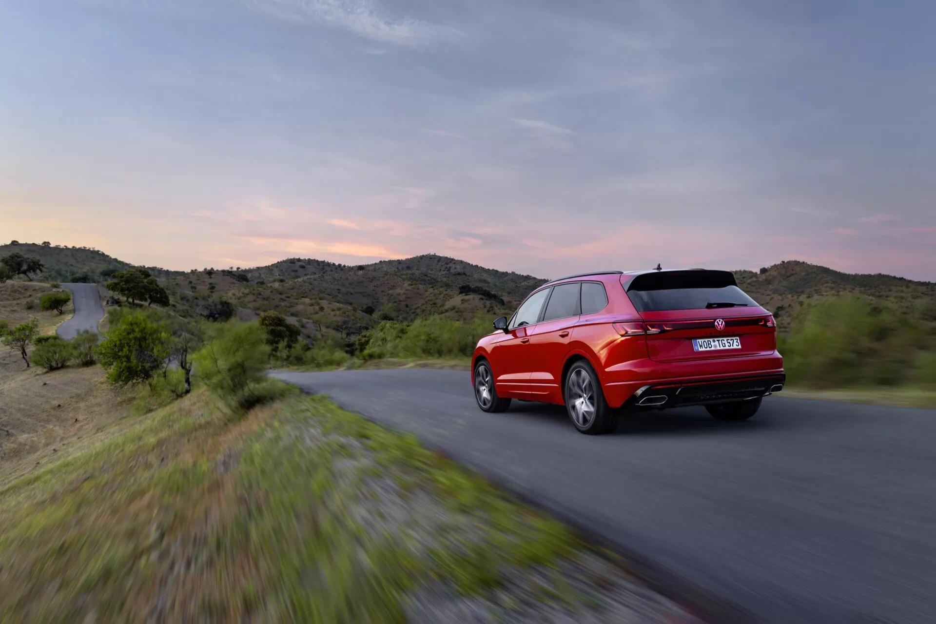 VW Touareg III Facelift 2023 v pohybe z pohľadu zo zadného trojštvrtečného uhla s LED zadnými svetlami a športovým kontúrom proti pozadiu večerného neba a kopcovitéj krajiny, zdôrazňujúci jeho červenú farbu, aerodynamický dizajn a výkonnostné prvky.