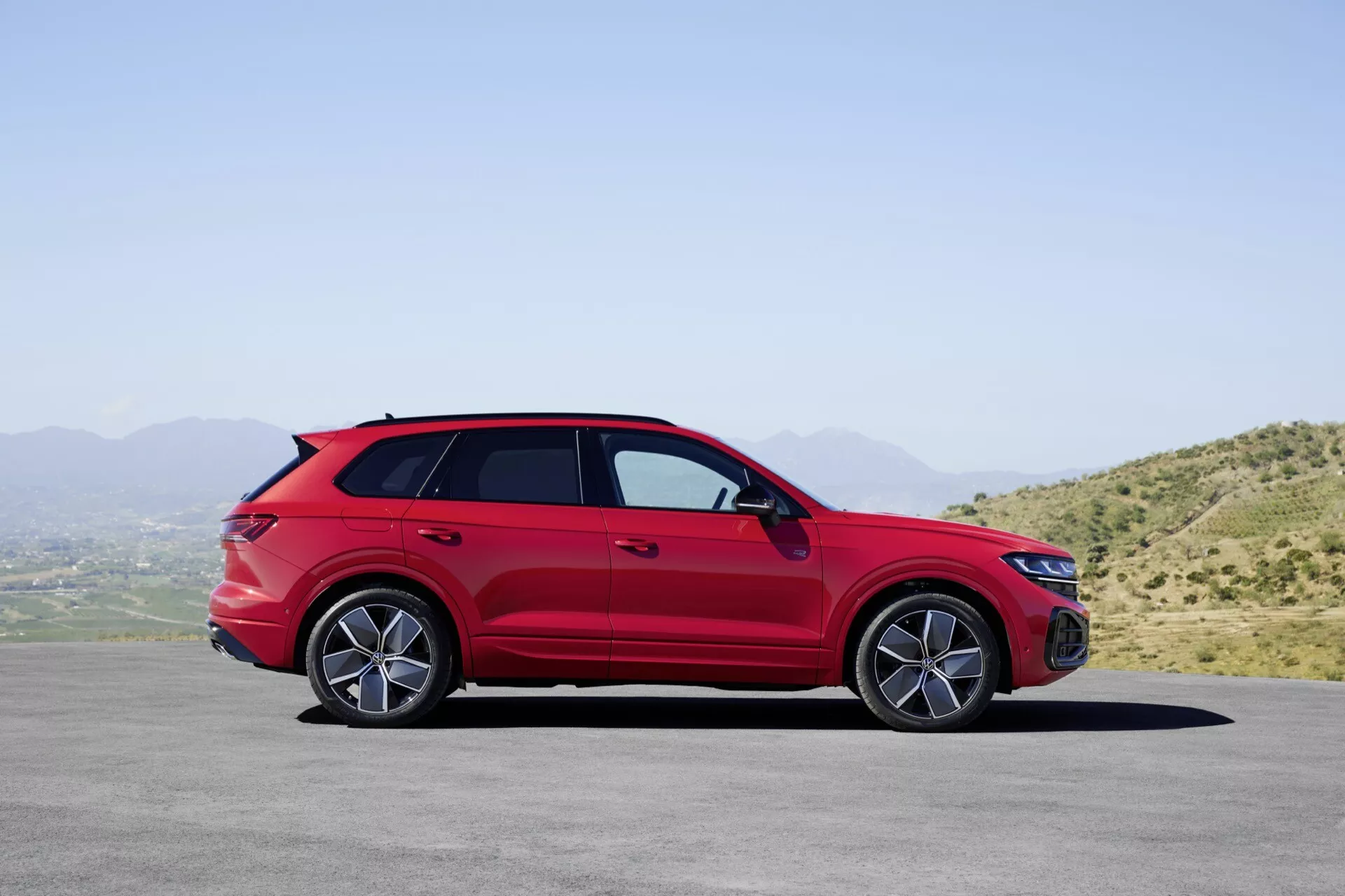 Model VW Touareg III po facelifte v červenej farbe zaparkovaný na rovnom povrchu s horskou krajinou a jasným nebom na pozadí, ukazujúci svoj bočný profil s detailným pohľadom na zliatinové kolesá, kľučky dverí, bočné zrkadlá s integrovanými smerovkami, strešné lyžiny, športové držanie tela v štýle kupé a tónované okná.