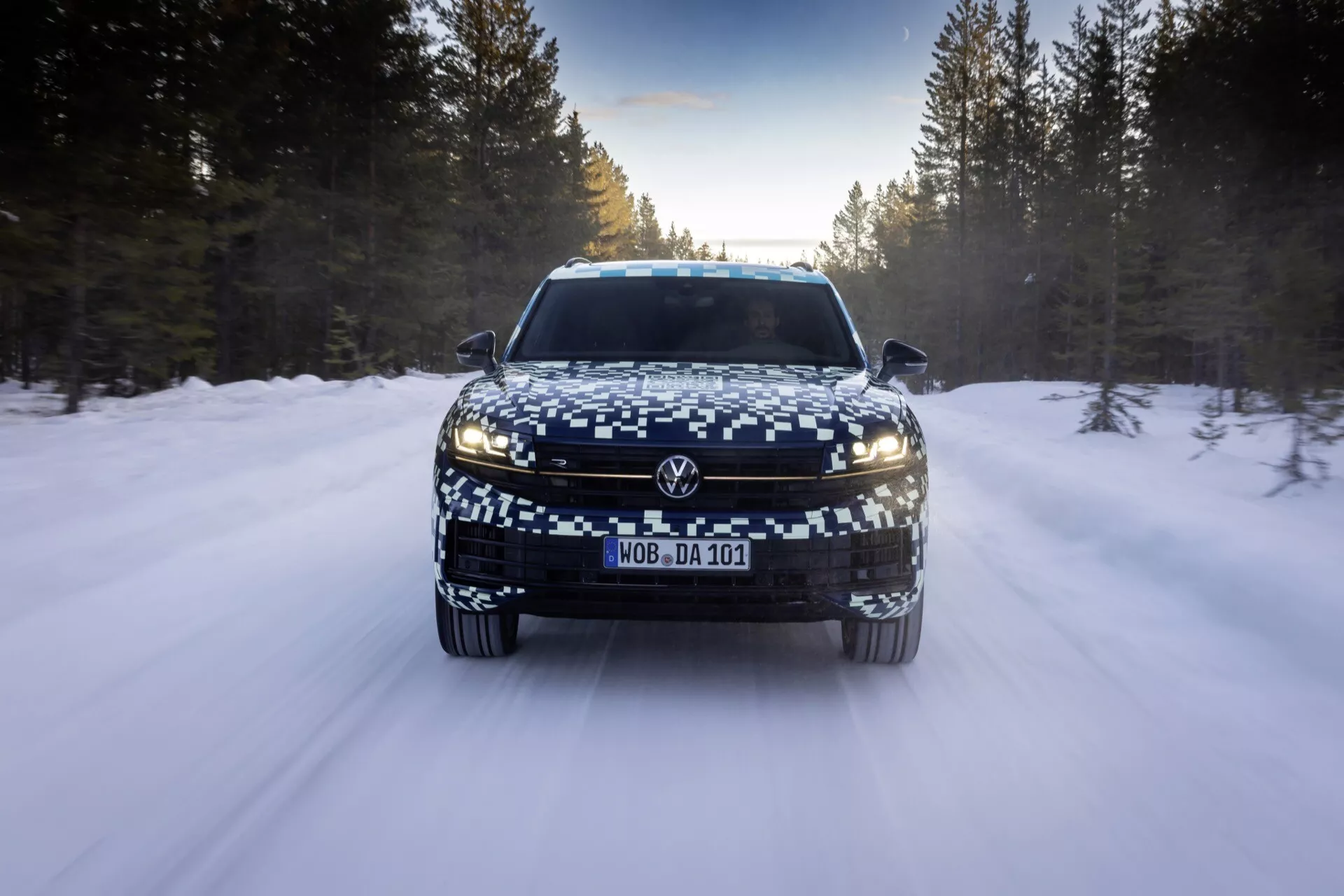 Predný pohľad na VW Touareg 2023 (III-Facelift) na ceste pokrytej snehom, predstavujúci LED svetlomety, širokú mriežku a maskovanú EČV, odrážajúci pokročilé osvetľovacie funkcie a funkcie pre zimnú jazdu.