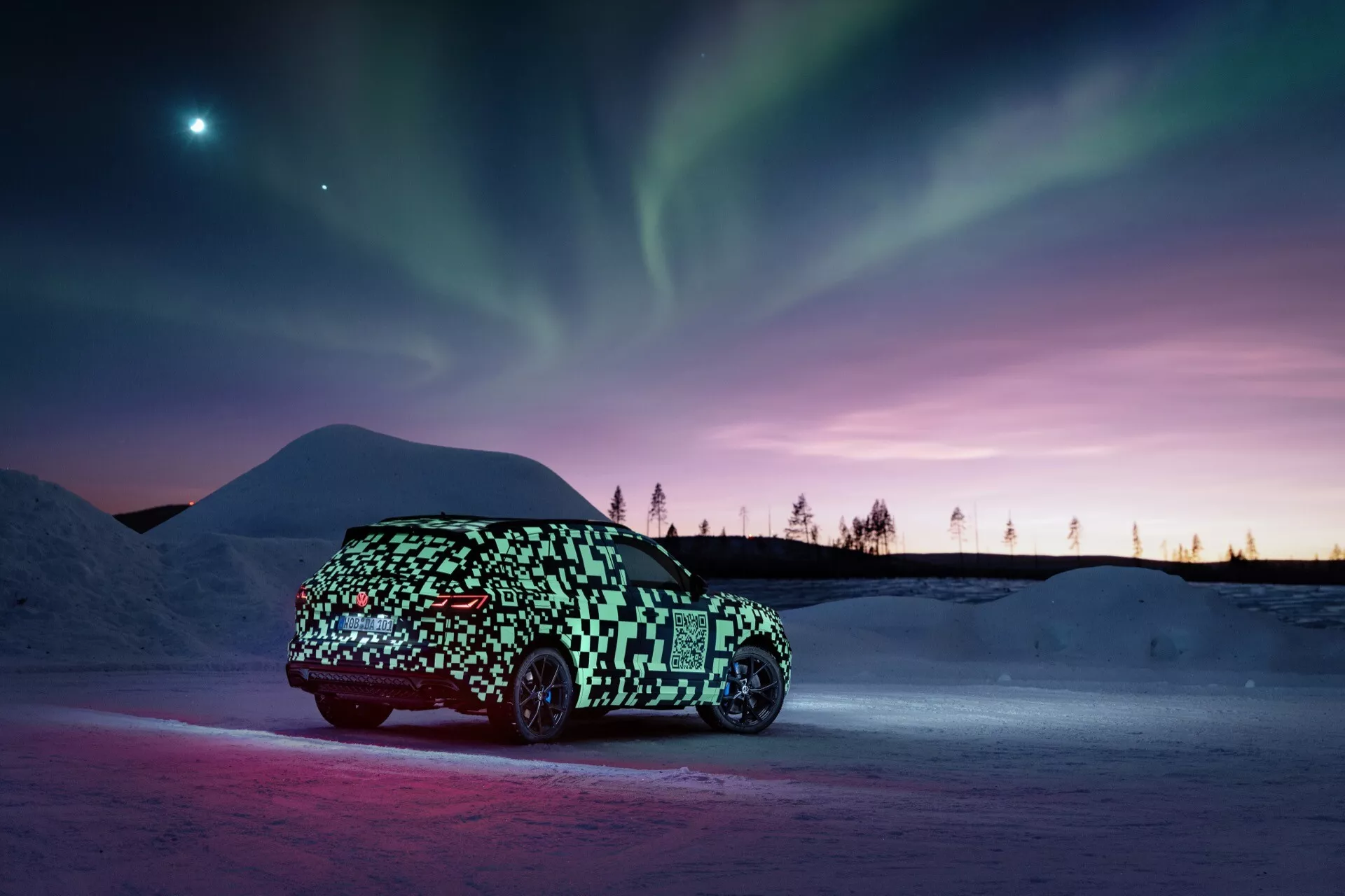 Prototyp VW Touareg (III-Facelift) 2023 v maskovacej folii zachytený z bočného profilu na snežnom podklade s ambientným osvetlením zdôrazňujúcim vozidlo proti pozadiu polárnej žiary a súmrakových odtieňov, naznačujúci elegantný, možno elektrický, dizajn hatchbacku alebo crossovera