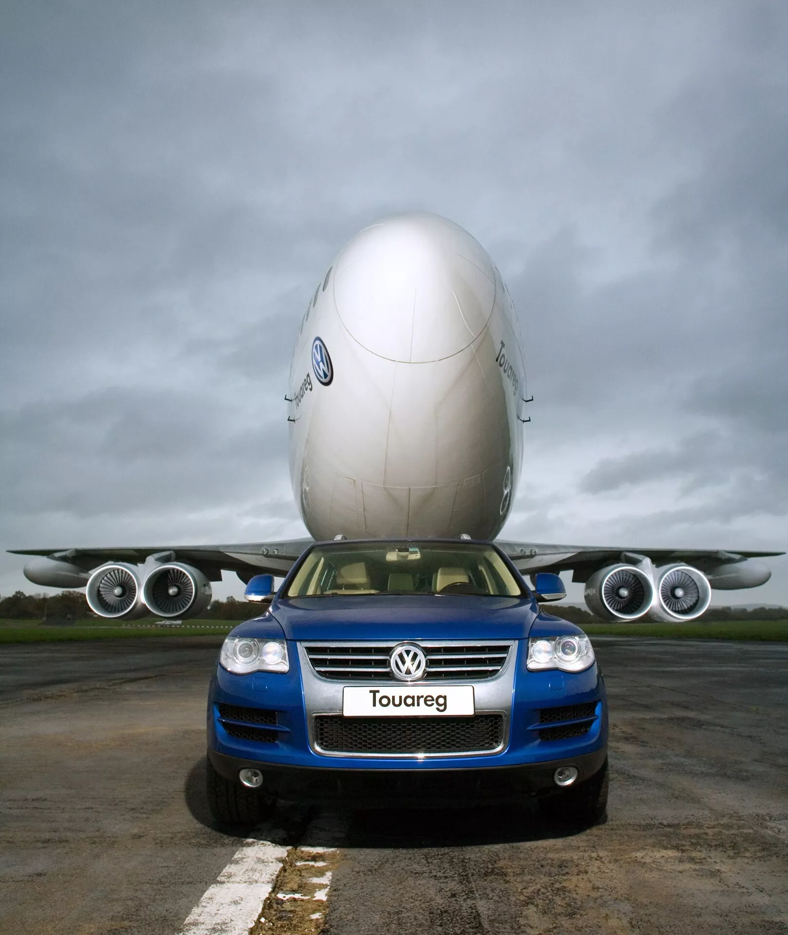 Modrý Volkswagen Touareg 2023 (III-Facelift) v popredí na ploche letiska, zachytený z nízkeho uhla zdôrazňujúceho jeho veľkosť a prednú časť, s mriežkou s logom, prednými svetlami, hmlistými svetlami a označením Touareg, s tvorivým pozadím lietadla zdajúceho sa byť odpočívajúcim na jeho streche proti zamračenej oblohe, zdôrazňujúc robustný dizajn a technologické funkcie vozidla