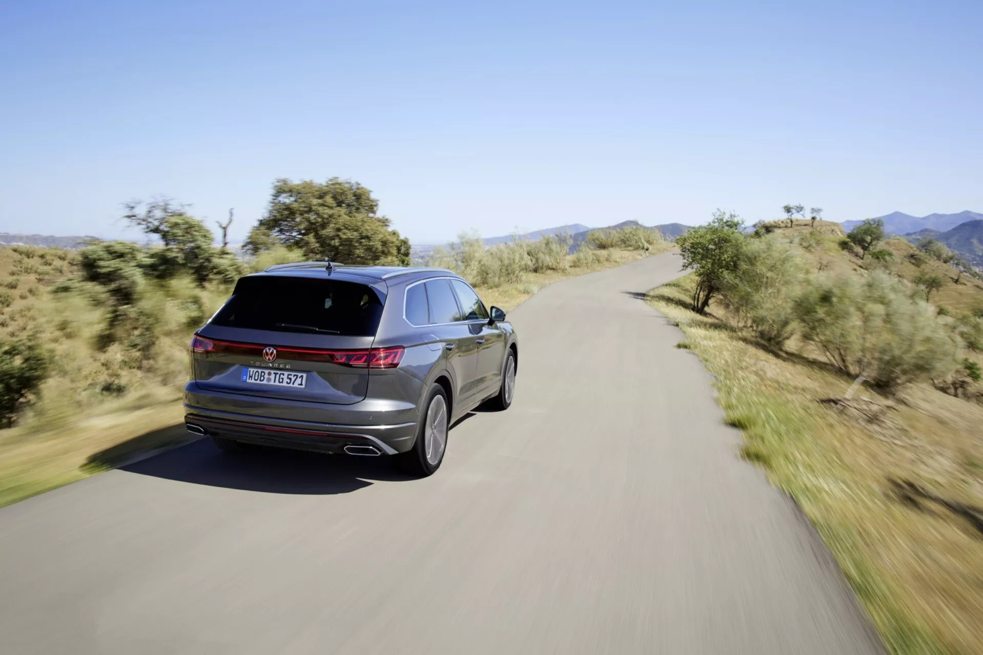 Zadný trojštvrťový pohľad na VW Touareg III-Facelift 2023 na scénickej ceste, zobrazuje LED zadné svetlá, charakteristické označenie, dvojité výfukové koncovky, zliatinové kolesá, elegantnú líniu strechy a lesklý povrch.