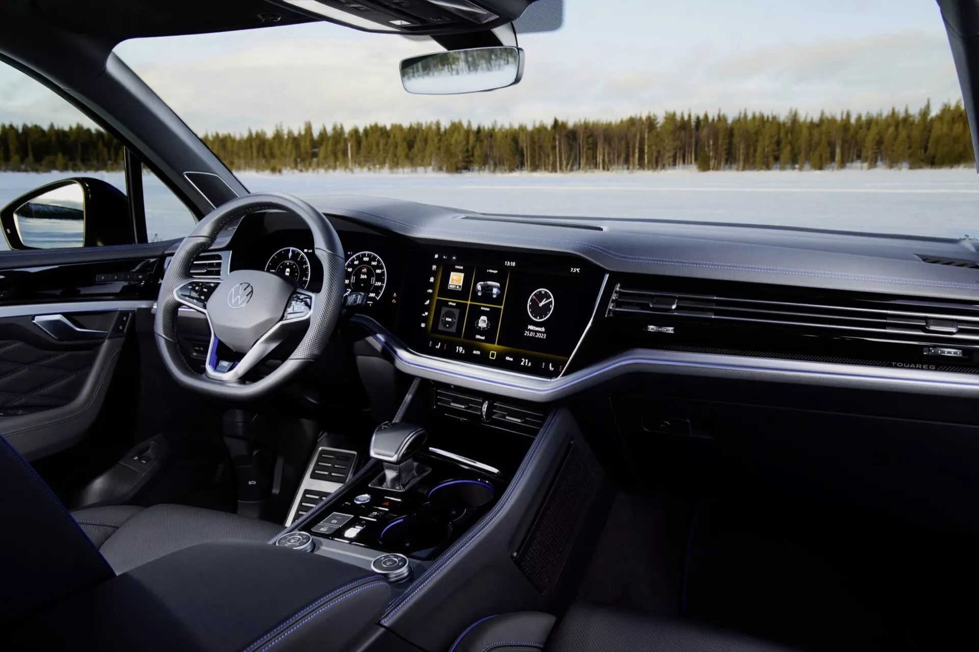 Interiér VW Touareg 2023 z pohľadu predného spolujazdca s čiernym koženým sedadlom, stredovou konzolou s pákou prevodovky, držiakmi na nápoje, ovládacími tlačidlami, dotykovým infotainment systémom, ventilačnými otvormi klimatizácie, digitálnym prístrojovým panelom a volantom s ovládaním, v pozadí zasnežená krajina cez čelné sklo, čo zdôrazňuje moderný luxus a technológie vozidla.