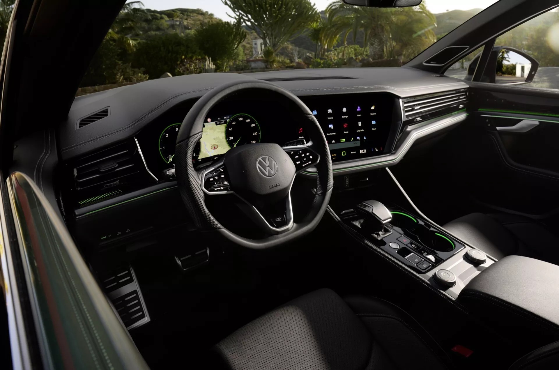 Interiér Volkswagen Touareg 2023 z pohľadu z zadného sedadla, predstavujúci panel dverí vodiča, prémiové ergonomické sedadlo vodiča, volant so znakom Volkswagen s namontovanými ovládacími prvkami, digitálny prístrojový panel a veľký dotykový informačno-zábavný systém s navigáciou, okrem minimalistického ovládania klímy a selektora automatickej prevodovky na stredovej konzole, čo odráža luxusné vlastnosti a pokročilú technológiu