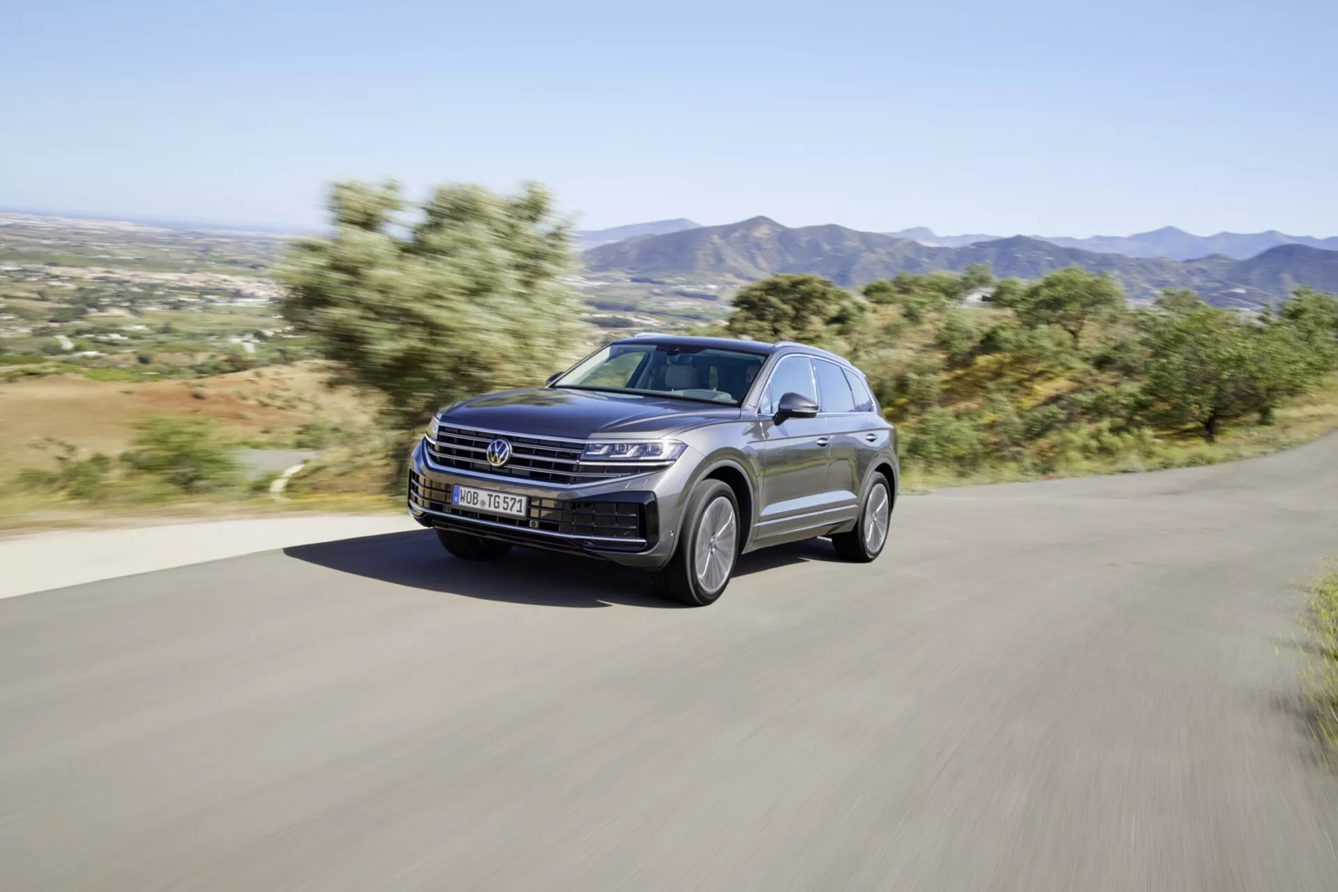 Model VW Touareg 2023 po facelifte dynamicky jazdiaci po spevnenej vidieckej ceste, prezentuje svoj moderný dizajn a výkonnostné prvky, vrátane LED svetlometov, luxusného metalického laku a štýlových zliatinových kolies, na pozadí horskej krajiny
