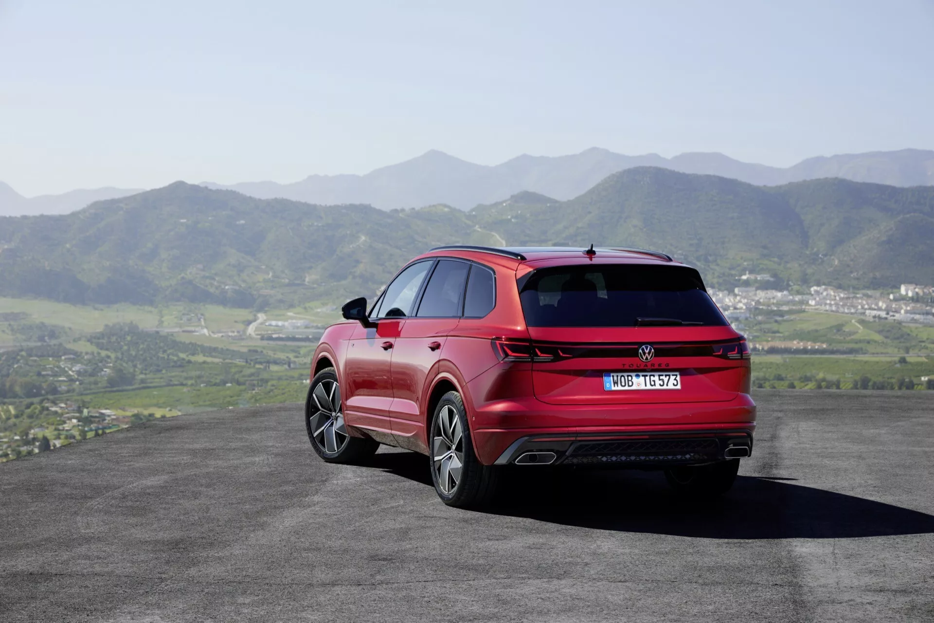 VW Touareg III Facelift 2023 v červenej farbe - pohľad zozadu a zo strany ukazuje LED zadné svetlá, dvojité výfuky, zliatinové kolesá a strieborné strešné lyžiny na pozadí valcovitých kopcov a čistého neba