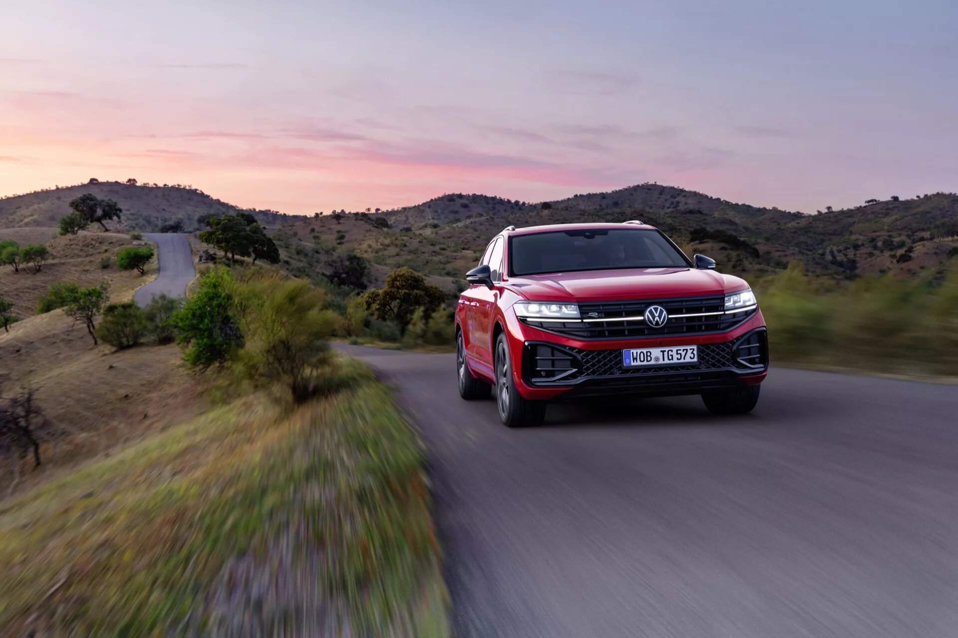 VW Touareg III-facelift 2023 v červenej farbe, zobrazujúci pohľad z prednej trojštvrťovej uhla s dôrazom na LED svetlomety, veľkú mriežku s emblémom, športovo aerodynamický dizajn, pokročilé zliatinové kolesá a funkcie ADAS v dynamickom pohybe na scénickej ceste za súmraku