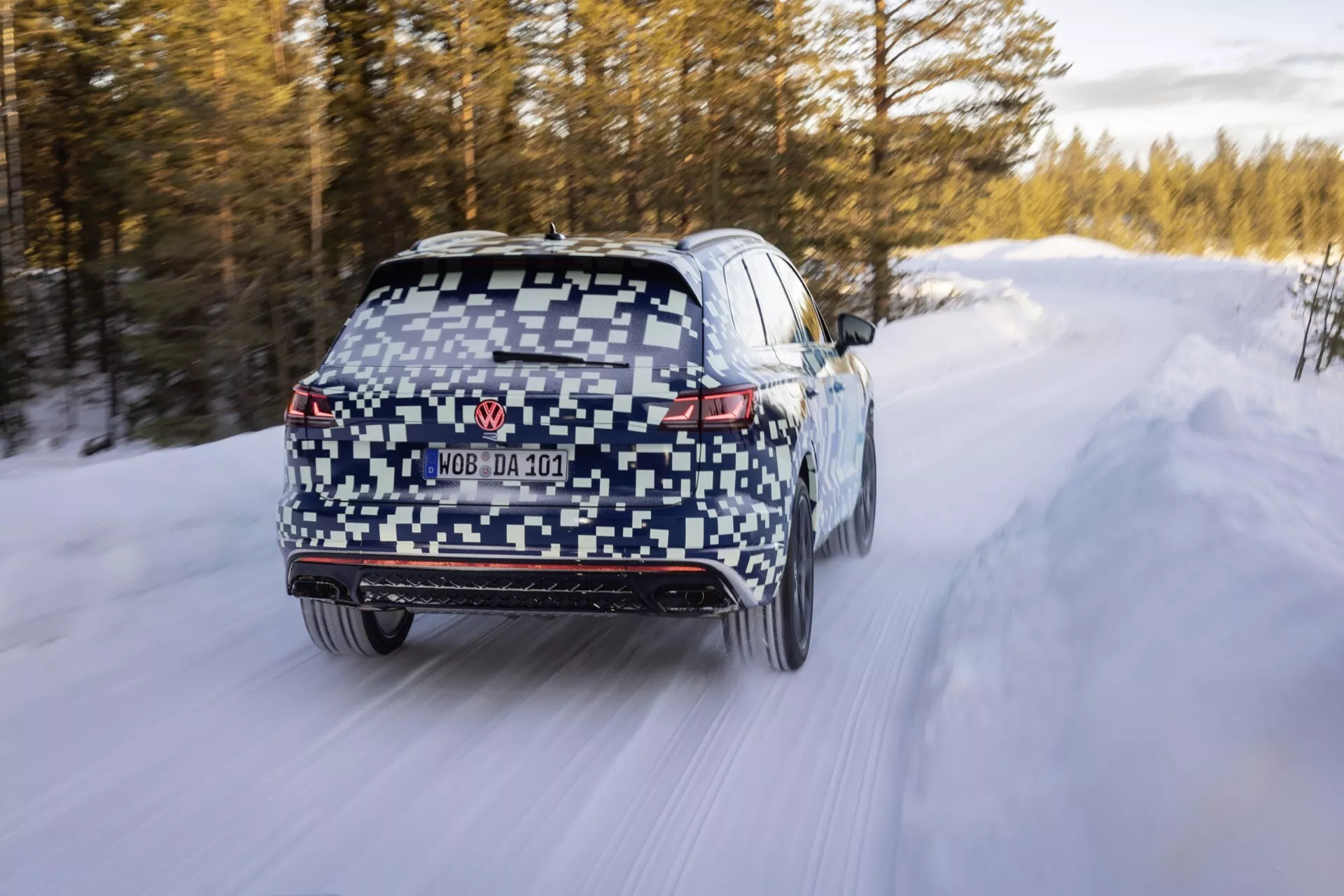 VW Touareg III Facelift 2023 v maskovaní idúci po zasneženej ceste so zadnými LED svetlami, zadným difúzorom, stieračom zadného okna, čiastočne skrytou EČV, dvojitými koncovkami výfuku, senzormi na zadnom nárazníku, rozostrením pohybu naznačujúcim pohyb, zimnými pneumatikami a potenciálnym pohonom všetkých kolies