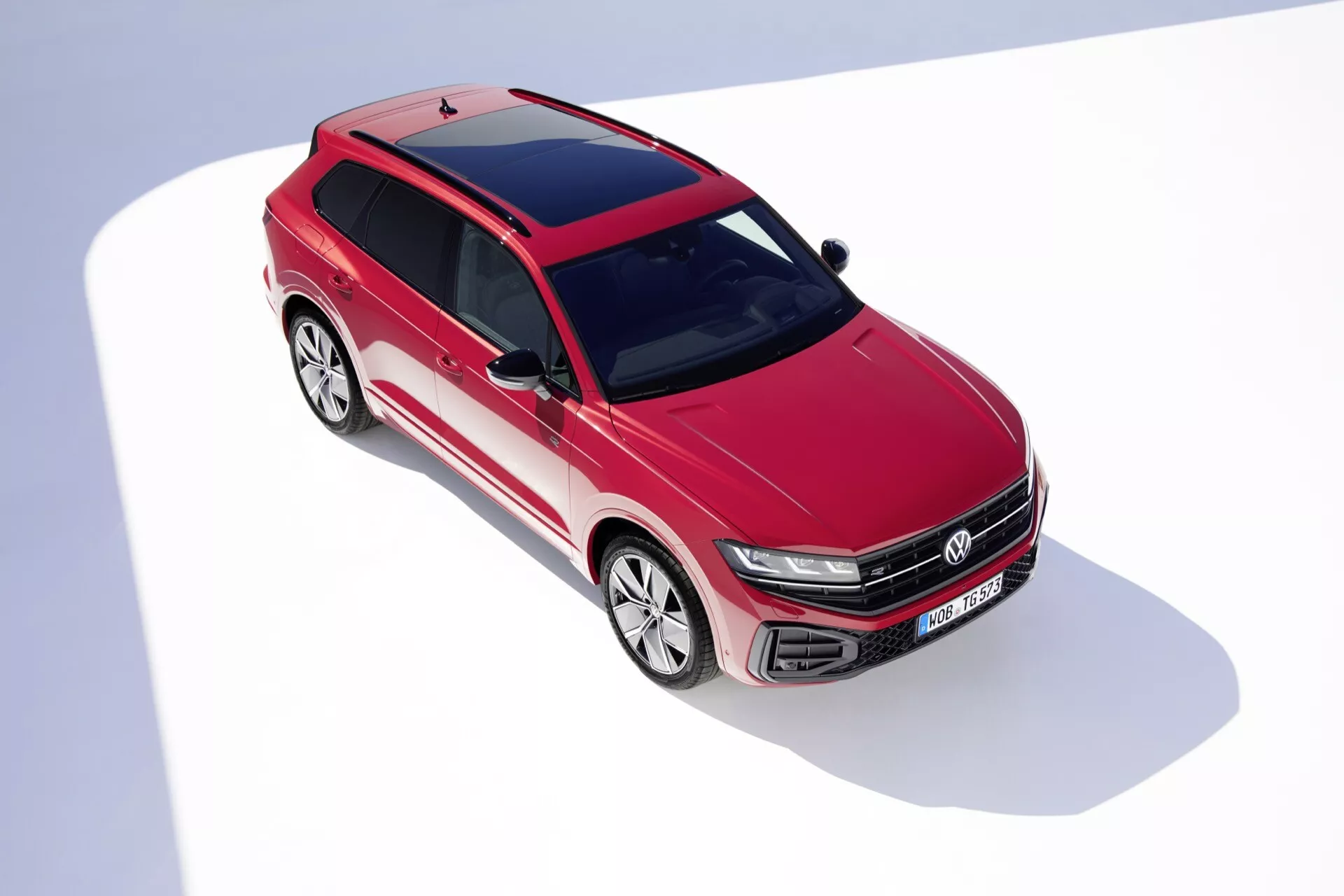 Vysokouhlý pohľad na prednú časť a bok vozidla VW Touareg III Facelift 2023 v červenej farbe, ktorý ukazuje dizajnové rysy vrátane mriežky s horizontálnymi lamelami a emblémom, LED predných svetlometov, aerodynamicky štýlovaného predného nárazníka s vetracími otvormi, tvarovaných liniek kapoty, rozsiahleho čelného skla, zliatinových kolies s viacerými ramenami, panoramatickej strechy a aerodynamických bočných zrkadiel na bielem pozadí
