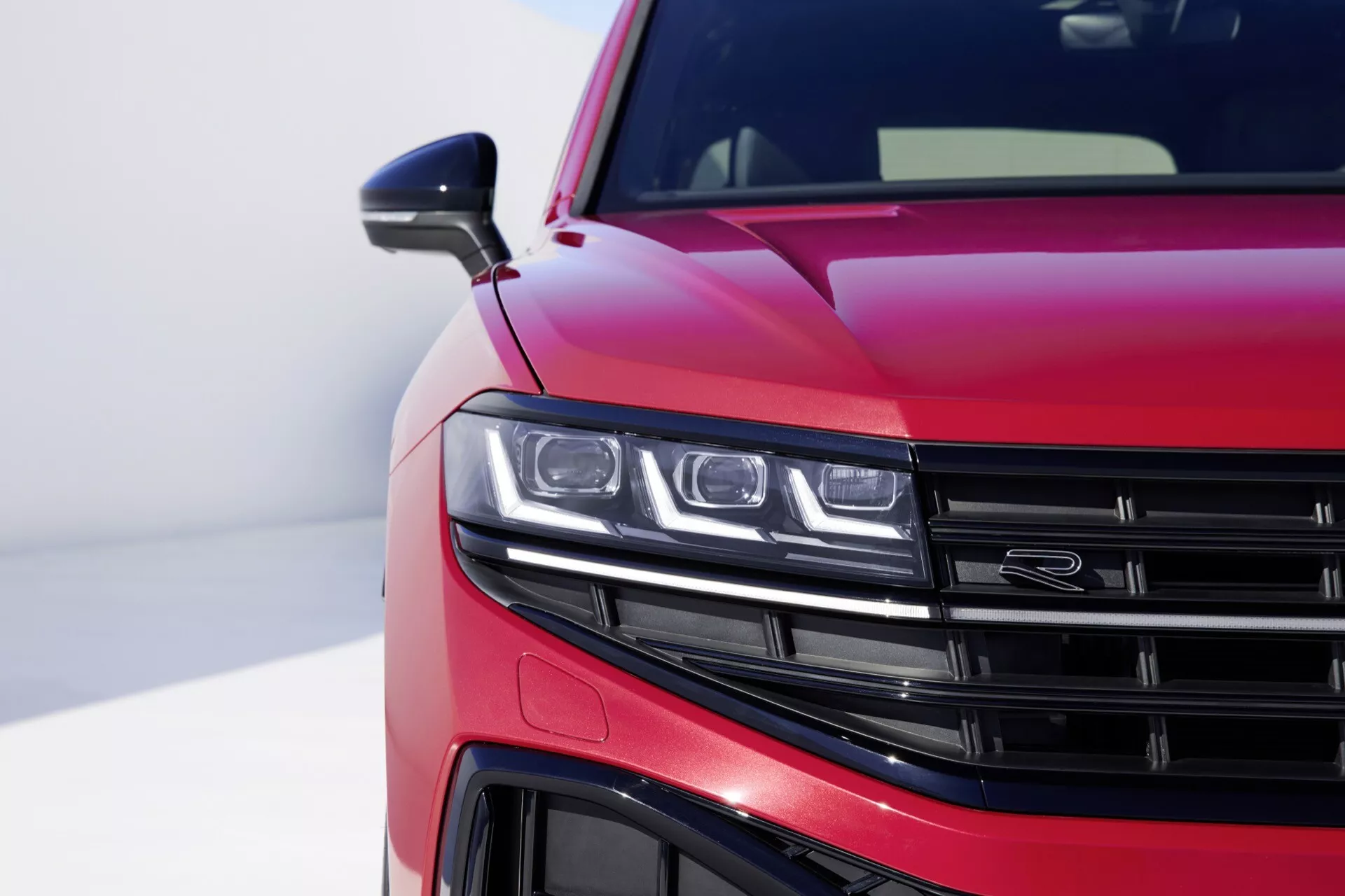 VW Touareg III-Facelift 2023 s lesklou metalickou červenou farbou zobrazujúci detailný pohľad na prednú ľavú štvrtinu, zobrazujúci súpravu LED svetlometov, časť mriežky s horizontálnymi lamelami, detailný pohľad na predný nárazník a aerodynamické línie na kapote proti rozmazanému pozadiu zdôrazňujúcim moderný dizajn a technologické vlastnosti auta.