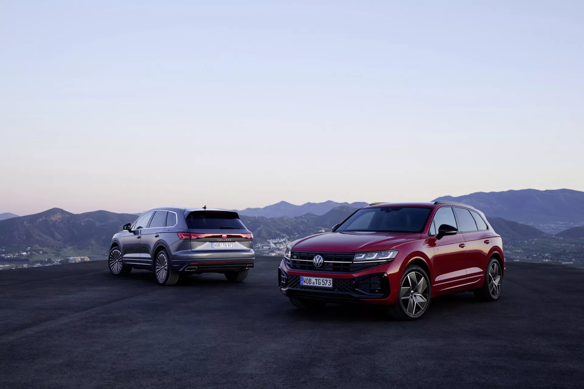 Modely Volkswagen Touareg (III-facelift) 2023 v červenej a striebornej farbe vonku s horskou panorámou a súmrakovou oblohou, prezentujúce predné a bočné profily s pokročilou LED svetelnou technológiou