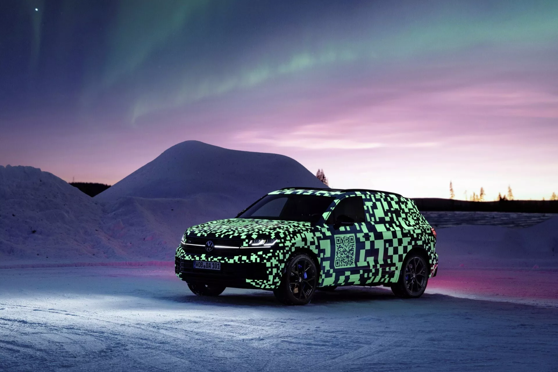 Model Volkswagen Touareg III facelift 2023 predstavený v zimnom súmraku, zobrazenie z pohľadu tri štvrtiny spredu so zameraním na LED svetlomety, prednú mriežku a maskovaciu fóliu na pozadí polárnej žiary a zasneženej krajiny