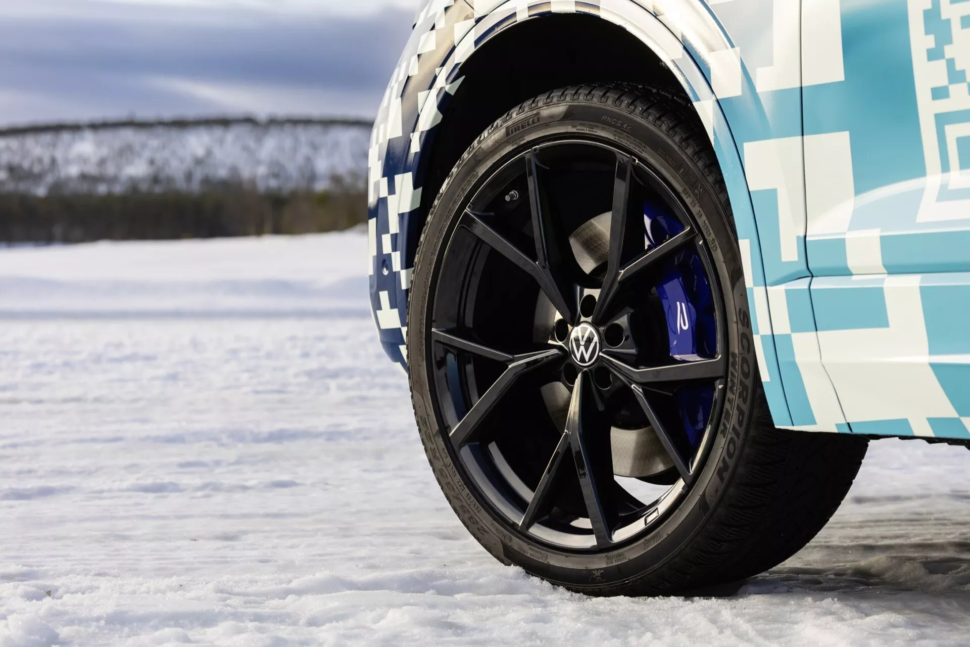 Detailný pohľad na predné koleso VW Touareg III Facelift 2023 s čiernym diskom s viacerými drážkami, logom Volkswagen a modrými brzdovými strmienami na snehovej ploche, predvádzajúce výkonnostné funkcie a zimné testovanie