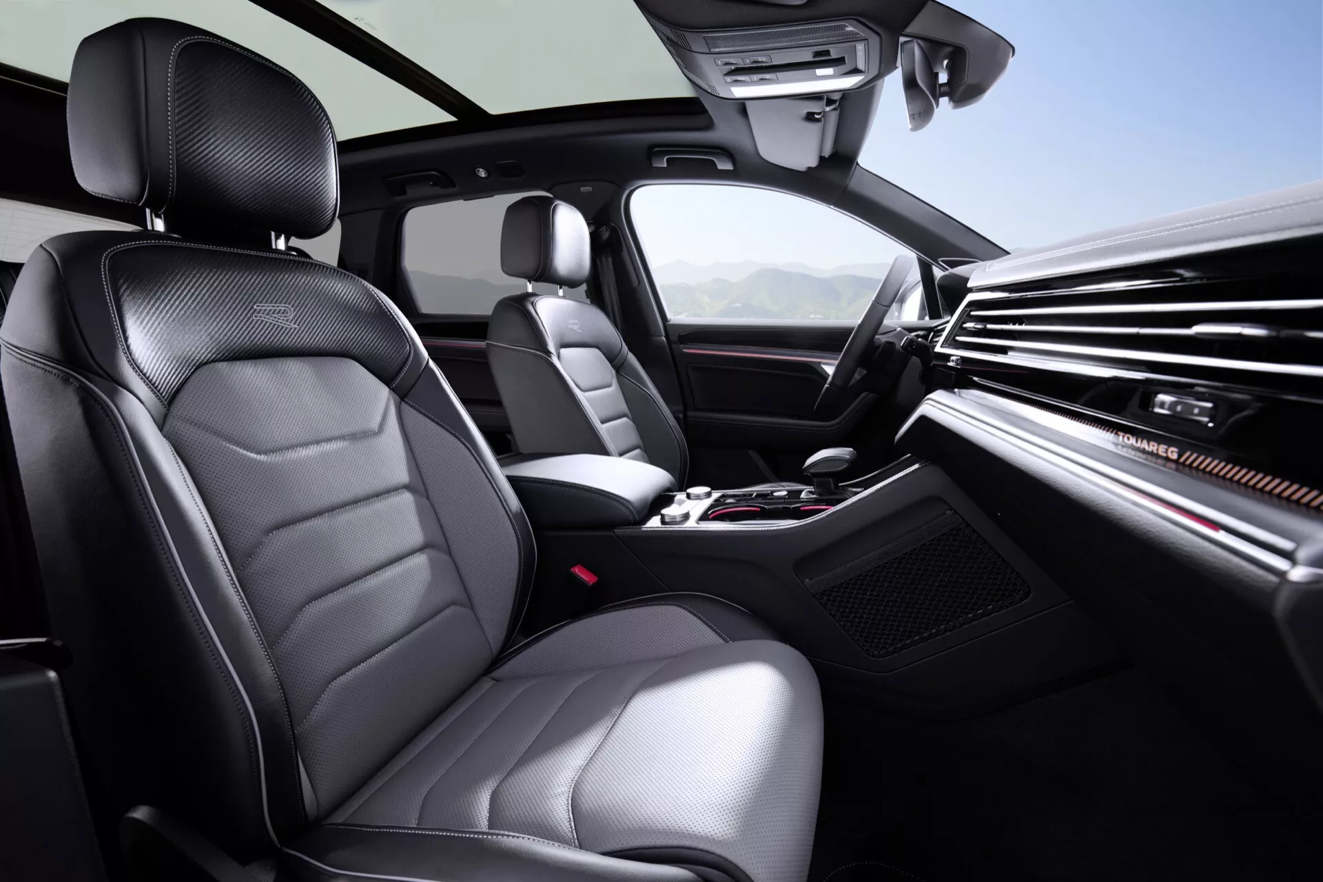 Interiérový pohľad na VW Touareg III Facelift 2023 zobrazujúci prednú časť pre cestujúceho a vodiča z perspektívy zadného sedadla, s prémiovými čalúnenými sedadlami s dekoračným šitím, rozsiahlym palubným doskou so štýlovými vetraními otvormi a centrálnym informačným displejom, zvýšenou konzolou uprostred s ovládaním klímy a médií a pákou automatickej prevodovky, obklopenou technológiou vrátane nastaviteľných sedadiel a ovládanie na volante, všetko osvetlené jasným interiérovým osvetlením pre čistú viditeľnosť.