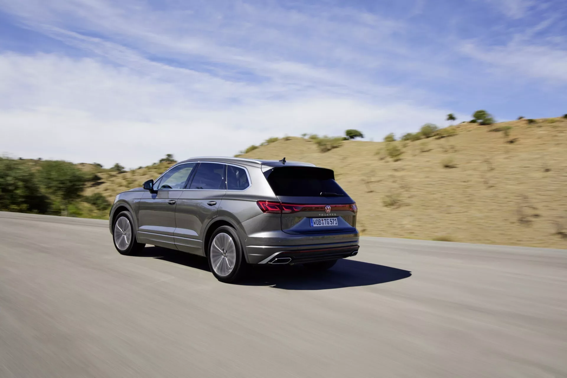 VW Touareg III facelift 2023 v pohybe na asfaltovej ceste, zachytený z zadného trojštvrtečného uhla ukazujúceho jeho elegantný dizajn, päťlúčové zliatinové kolesá, metalický finiš, LED zadné svetlá, integrované výfuky a športovú estetiku proti suché, kopcovitej krajine pod jasne slnečnou oblohou