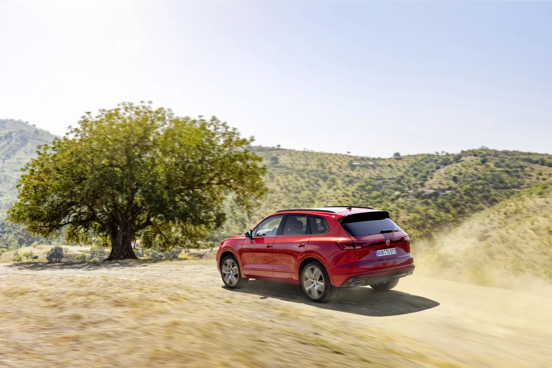 Červené SUV Volkswagen Touareg III Facelift 2023 ukazujúce schopnosti v teréne na neudržiavanej ceste, zozadu z trojštvrťového uhla zdôrazňujúci jeho dynamický dizajn, disky kolies, bočné dvere, bočné a zadné okná, s prachom naznačujúcim pohyb a výkonnostné charakteristiky ako výkon alebo pohon 4x4, v prirodzenom vonkajšom prostredí naznačujúcom dobrodružstvo a všestrannosť v teréne.