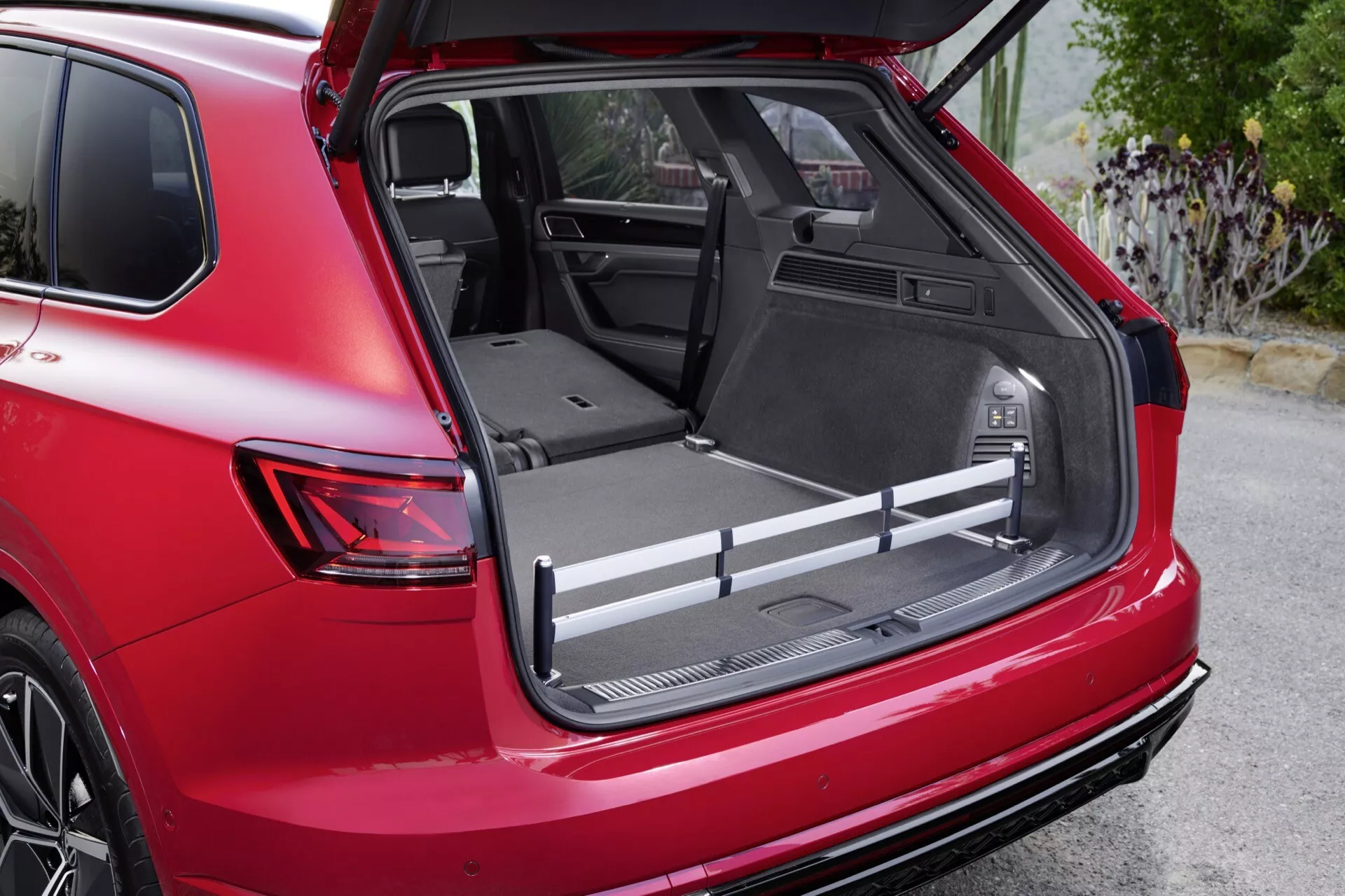 Detailný obrázok Volkswagen Touareg III po facelifte z roku 2023 ukazujúci priestranný nákladný priestor s plochou nákladovou plochou, ochranným obkladom a integrovanými úložnými priestormi z pohľadu od zadnej pravej strany pod uhlom tri štvrtiny, s 12V zásuvkou, nastaviteľnými nákladnými tyčami a čiastočne sklopenými zadnými sedadlami pre rozšírenie priestoru, zdôraznený červenou vonkajšou farbou vozidla a športovým zadným spojlerom.